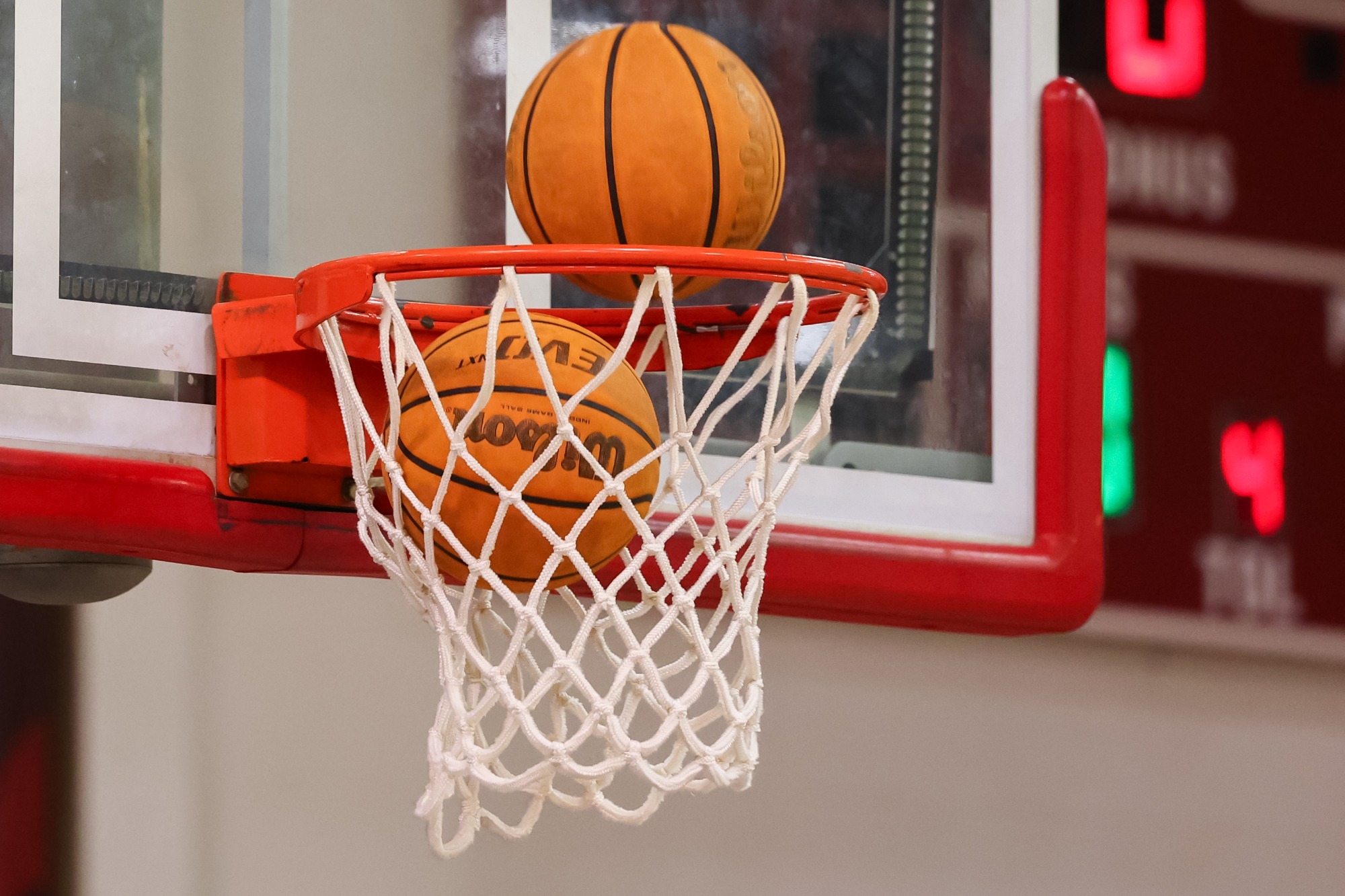 Close-up of a basketball balanced on the rim as another ball passes through the net during warmups or practice. The backboard and hoop fill the frame, with the scoreboard softly blurred in the background of the gymnasium