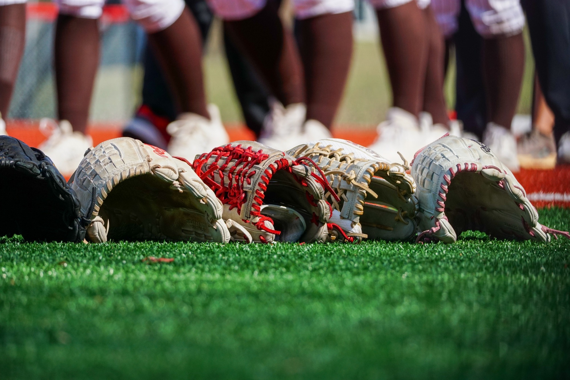Several softball gloves lie in a row on the grass in the foreground, with players’ legs and cleats visible in the background. The gloves feature white, red, tan, and black leather with contrasting laces