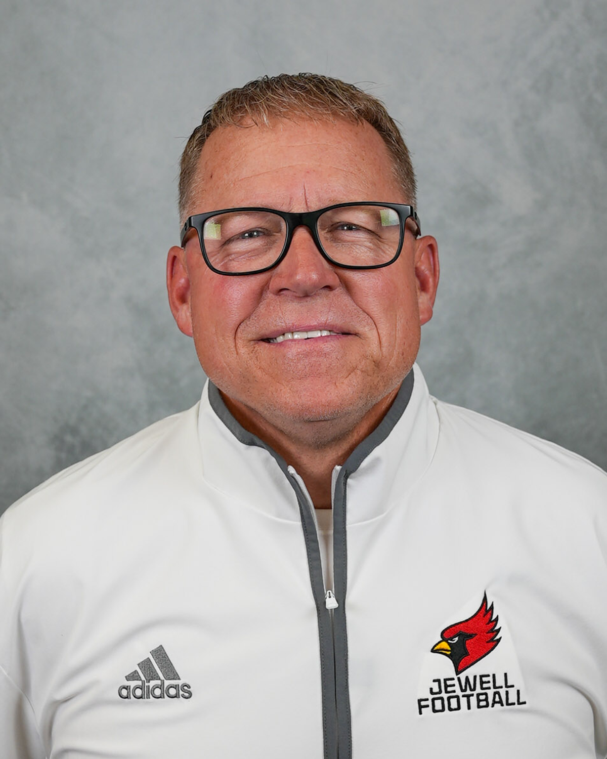 Headshot of William Jewell football coach Jason Ambroson wearing a white Jewell Football pullover and glasses in front of a gray background