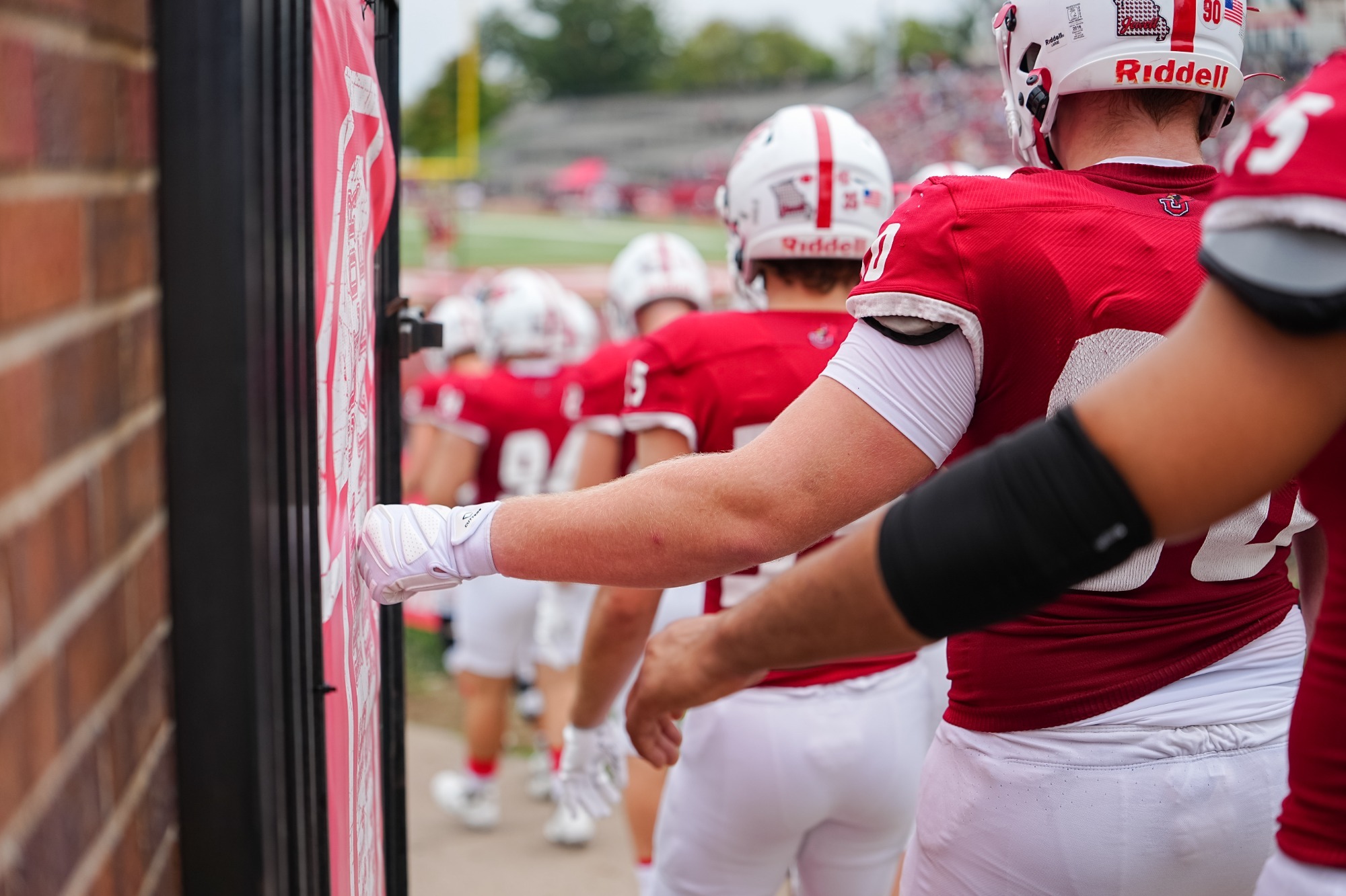 William Jewell football players in red uniforms walk onto the field through a stadium tunnel, with one player in the foreground touching a wall graphic