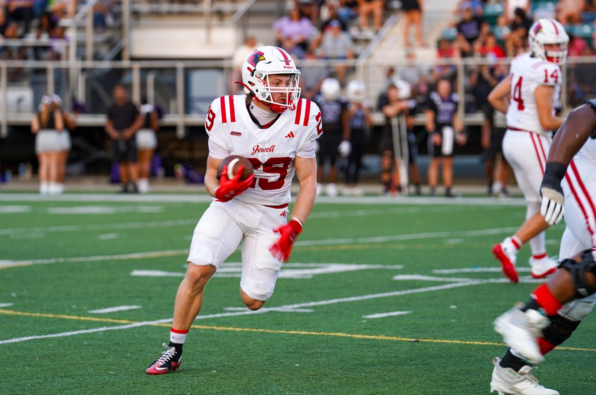 Hunter Bailey wearing a white uniform and helmet, holding a football and running during a game with a stadium in the background