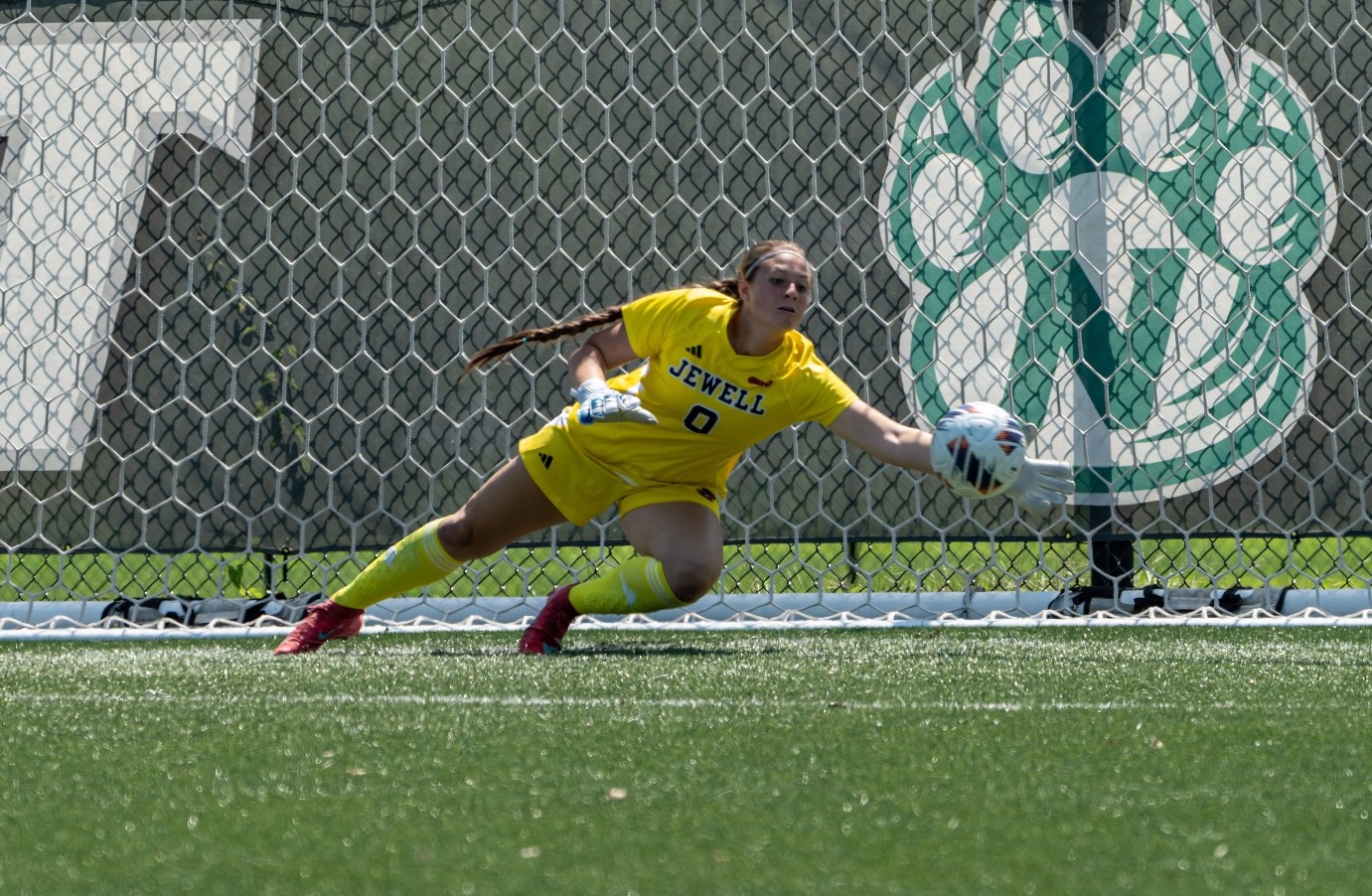 Goal keeper in yellow jersey and shorts dives to her left to stop of soccer ball with her outstretch arm
