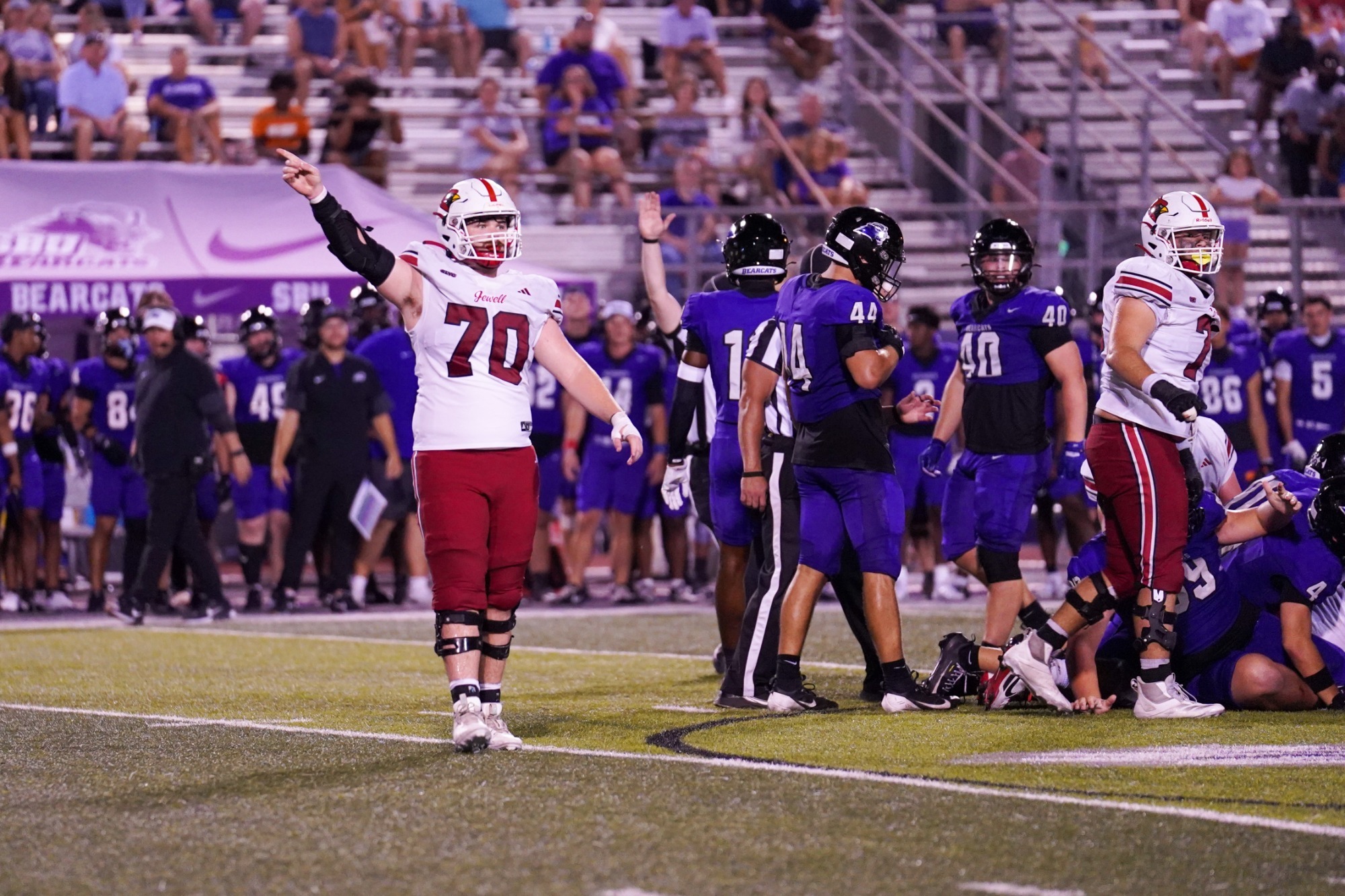Zach Barnhouse standing at midfield in a white football jersey, red pants, and white helmet pointing to the left signaling a first down with a stadium in the background and Southwest Baptist players in the pile to the right