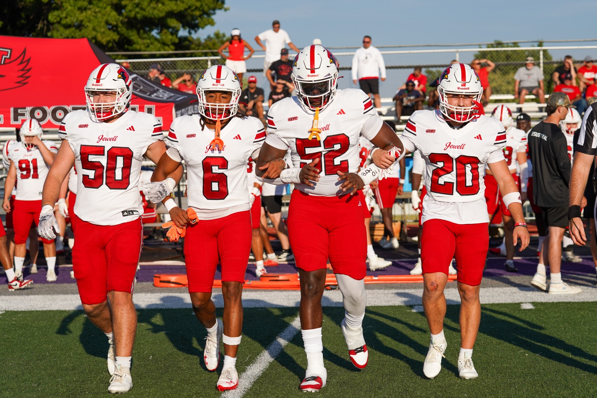 The four football captains, wearing red pants, white jerseys, and white helmets, walk arm in arm 