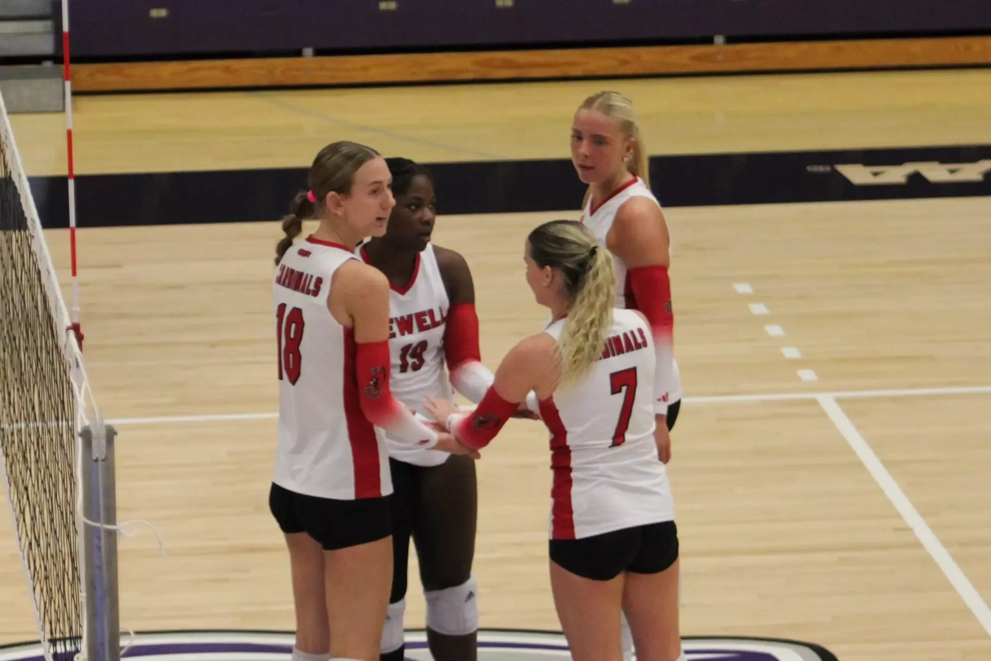 Kali Adamec, Brianna Witthar, Alexis Frazier, and Macy Barlett standing in a huddle while wearing white volleyball jerseys