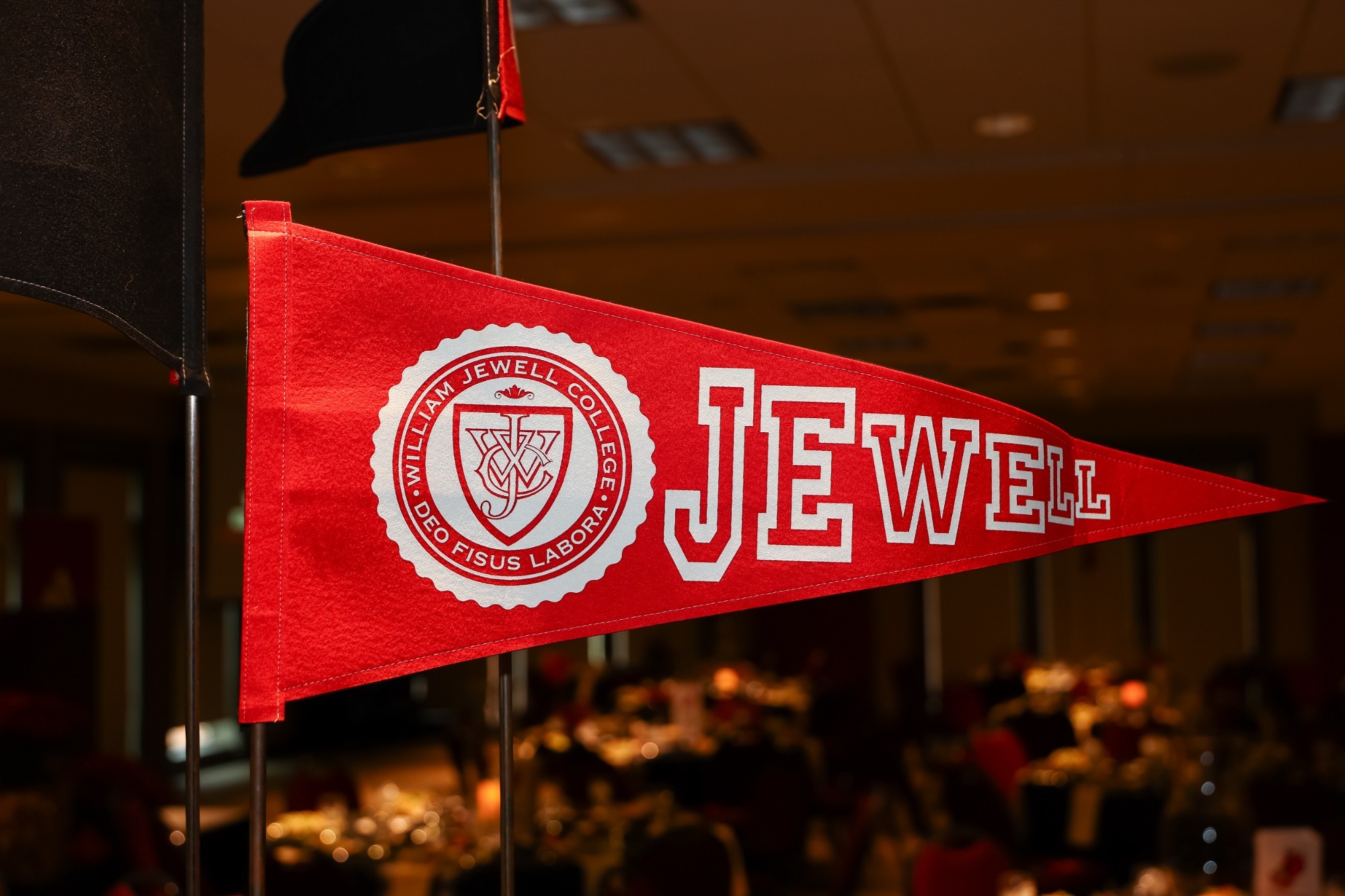 Red pennant banner with the William Jewell seal and the Jewell written in white with banquet tables blurred in the background