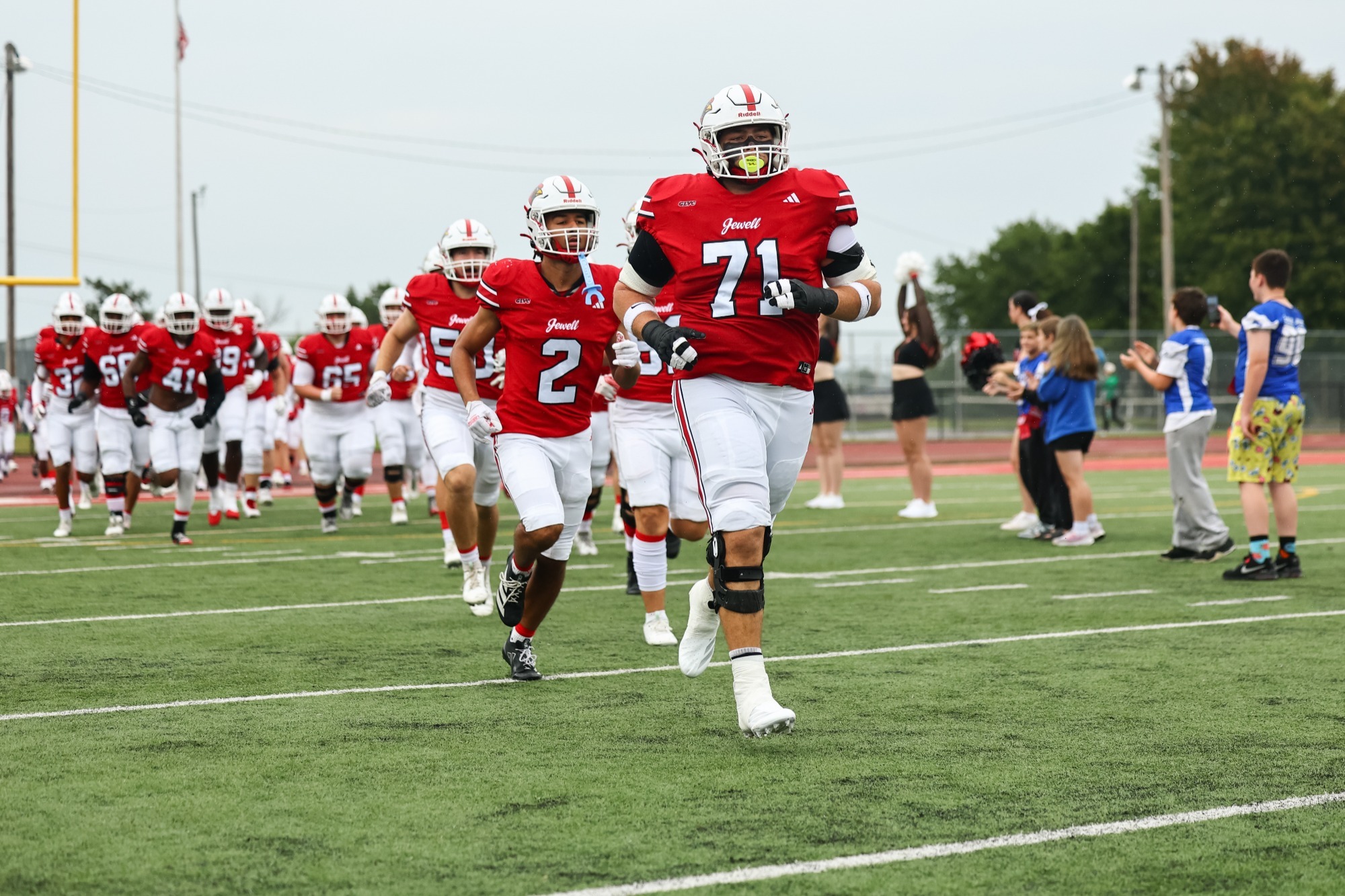 Members of the William Jewell football team run out onto the field, wearing white helmets, red uniforms, and white pants with youth football players in blue jerseys in the background