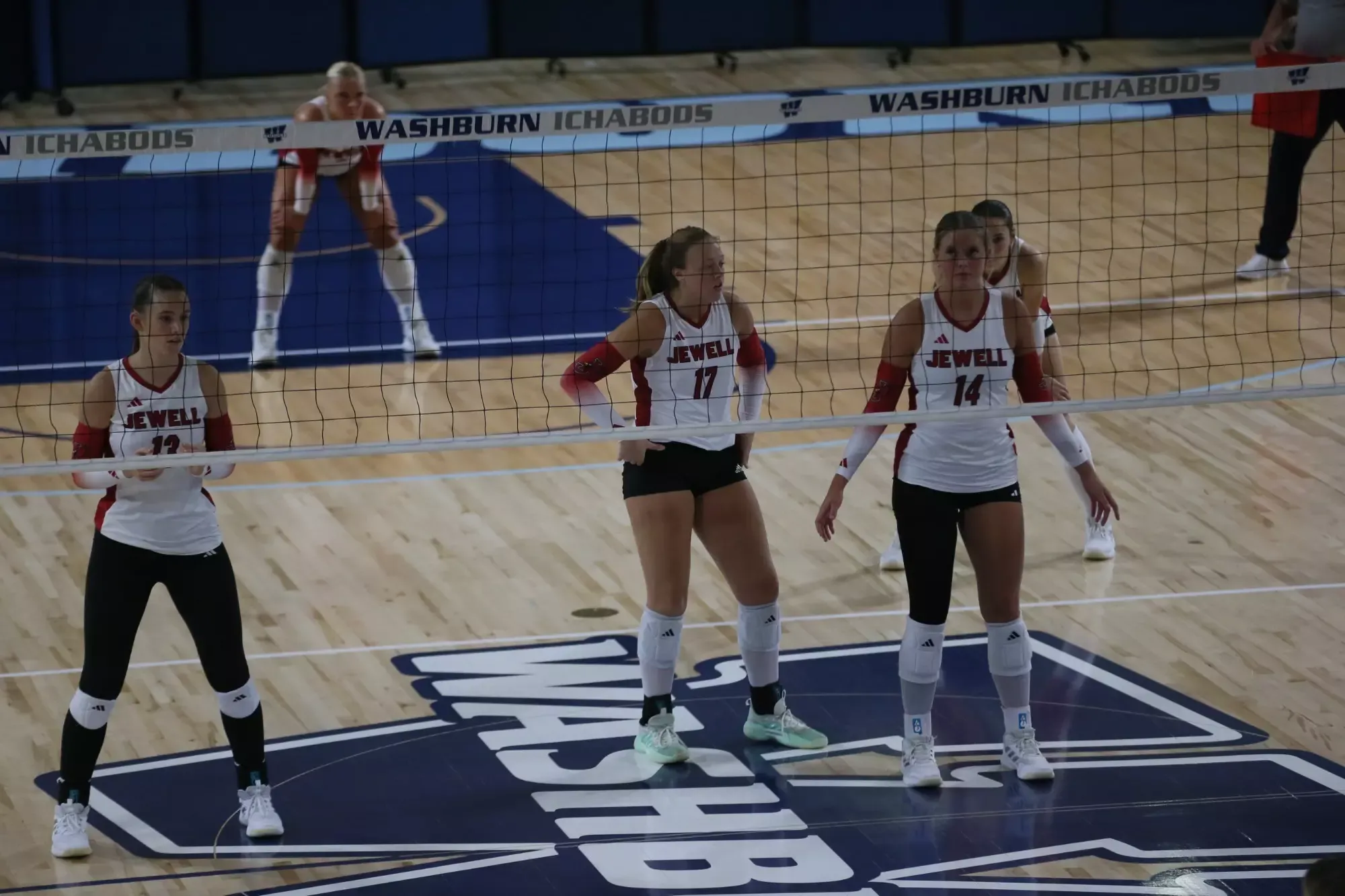 Three volleyball players wearing a white Jewell uniform, standing behind a blurred net in the foreground