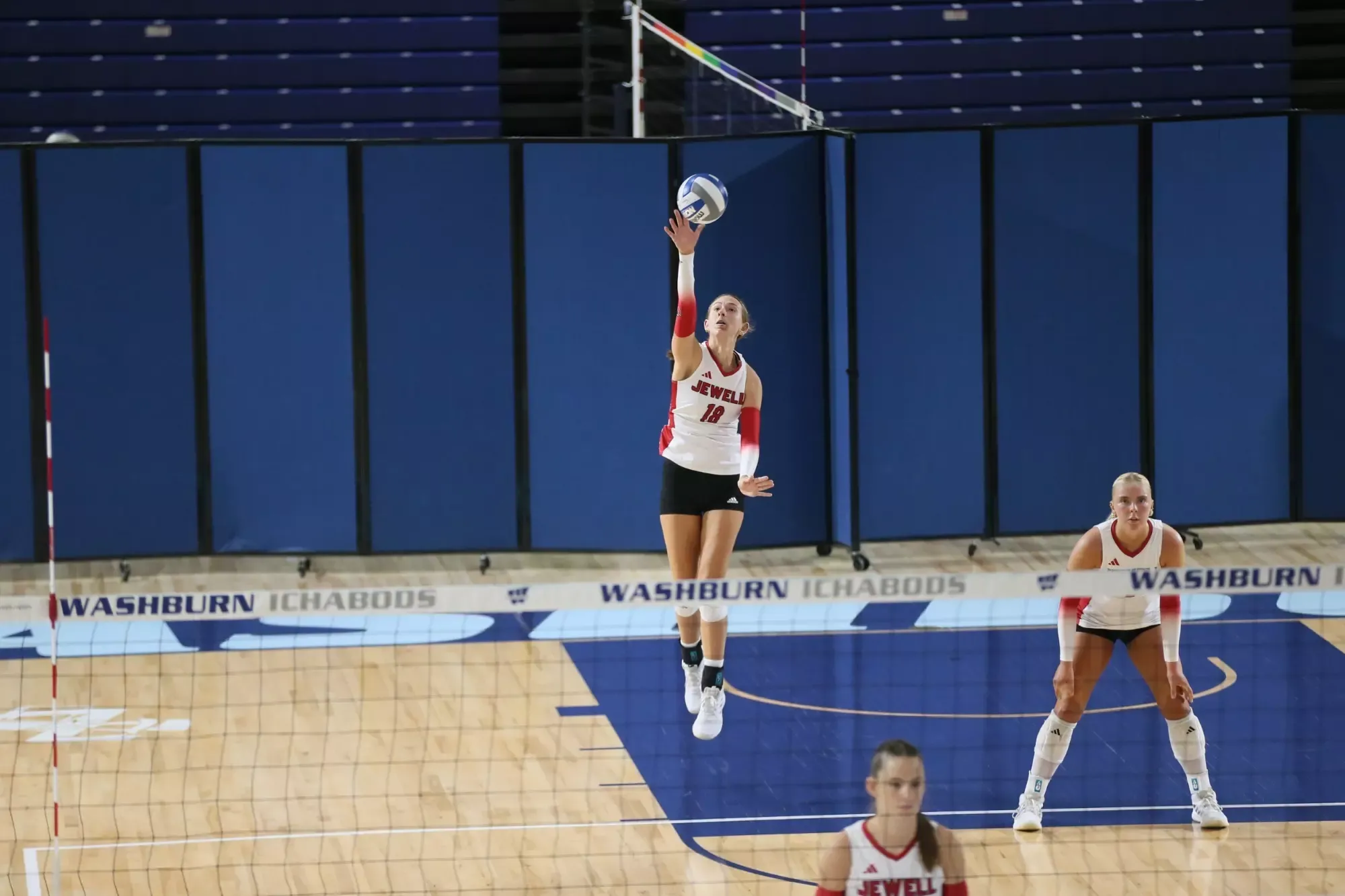 Kali Ademec jumping to serve a volleyball, wearing a white uniform with a blurred volleyball net in the foreground