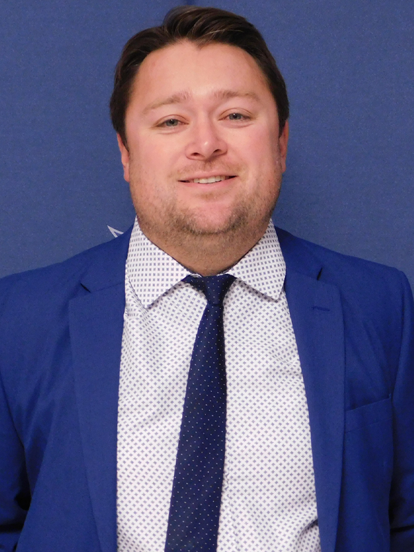 Assistant Men's Basketball Coach Kirk Fallon in a blue suit with a blue tie, in front of a blue Lackawanna College background