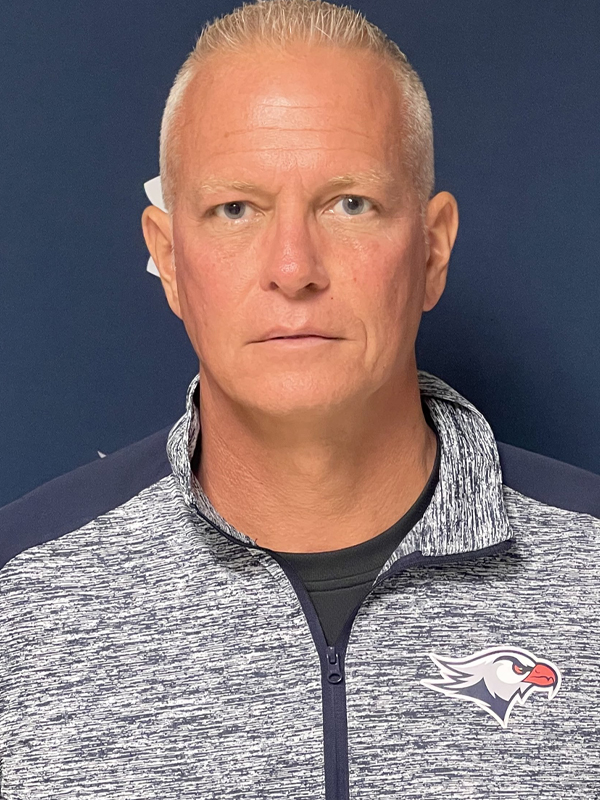 Assistant Women's Basketball Coach Doug Miller, standing in front of a blue background with the Falcon logo on it, in a grey and blue quarter-zip pullover with the Falcon logo on the left chest.