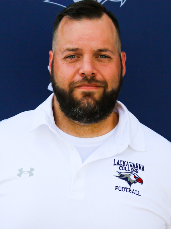 Assistant Football Coach Bill Reiss In A White Shirt Standing In Front Of A Blue Lackawanna College Falcons Backdrop