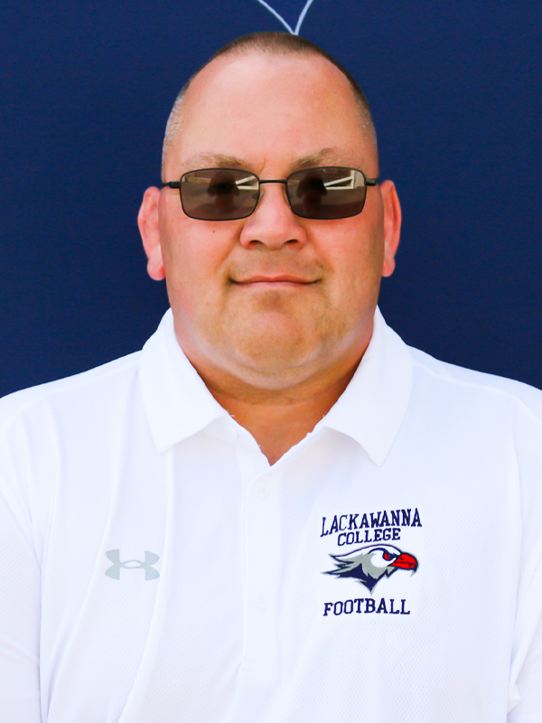 Assistant Football Coach Kevin Roginski In A White Shirt Standing In Front Of A Blue Lackawanna College Falcons Background