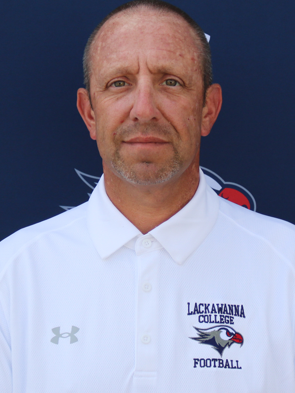 Assistant Football Coach Michael Behr Standing In A White Shirt In Front Of The Blue Lackawanna College Falcon Backdrop