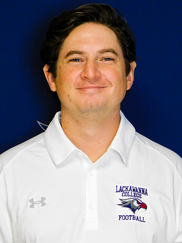 Athletic Trainer Frank Mroz In A White Shirt Standing In Front Of A Blue Lackawanna College Falcons Background.