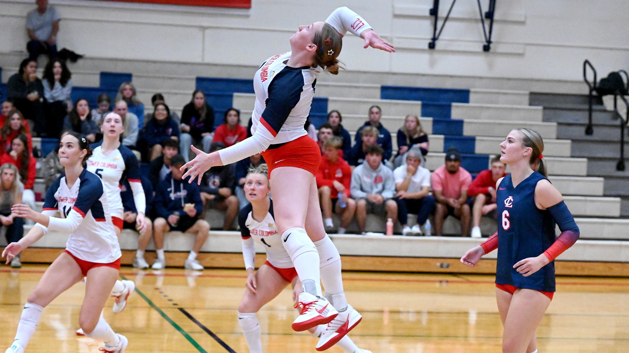 A Lower Columbia College volleyball player leaps mid-air to hit a volleyball above the court during a indoor match, surrounded by teammates and spectators.