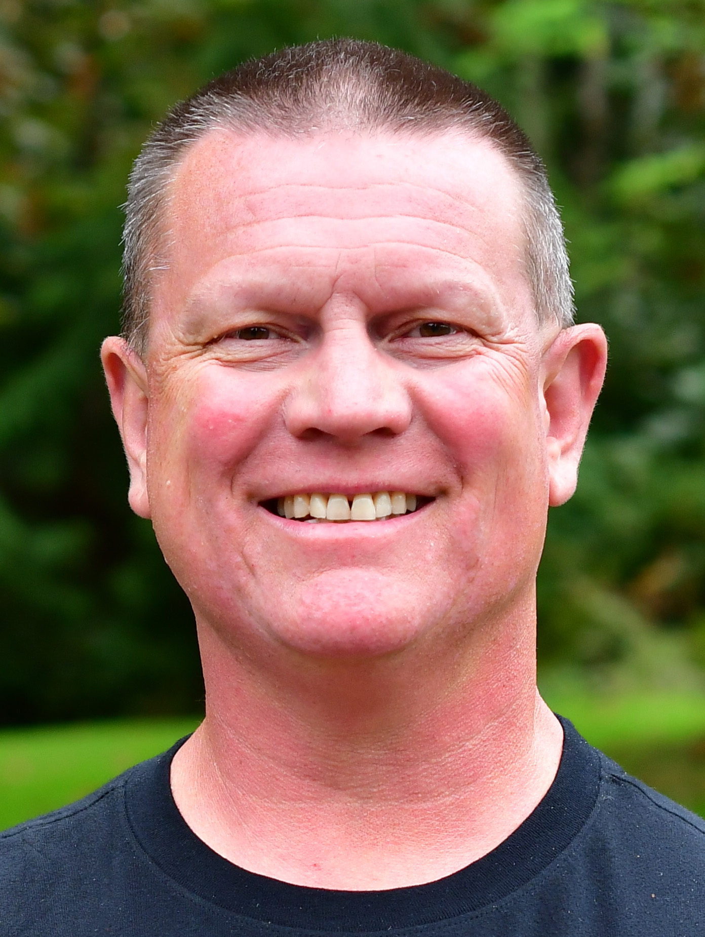 Scott Campbell stands outside in front of a green background, smiling.