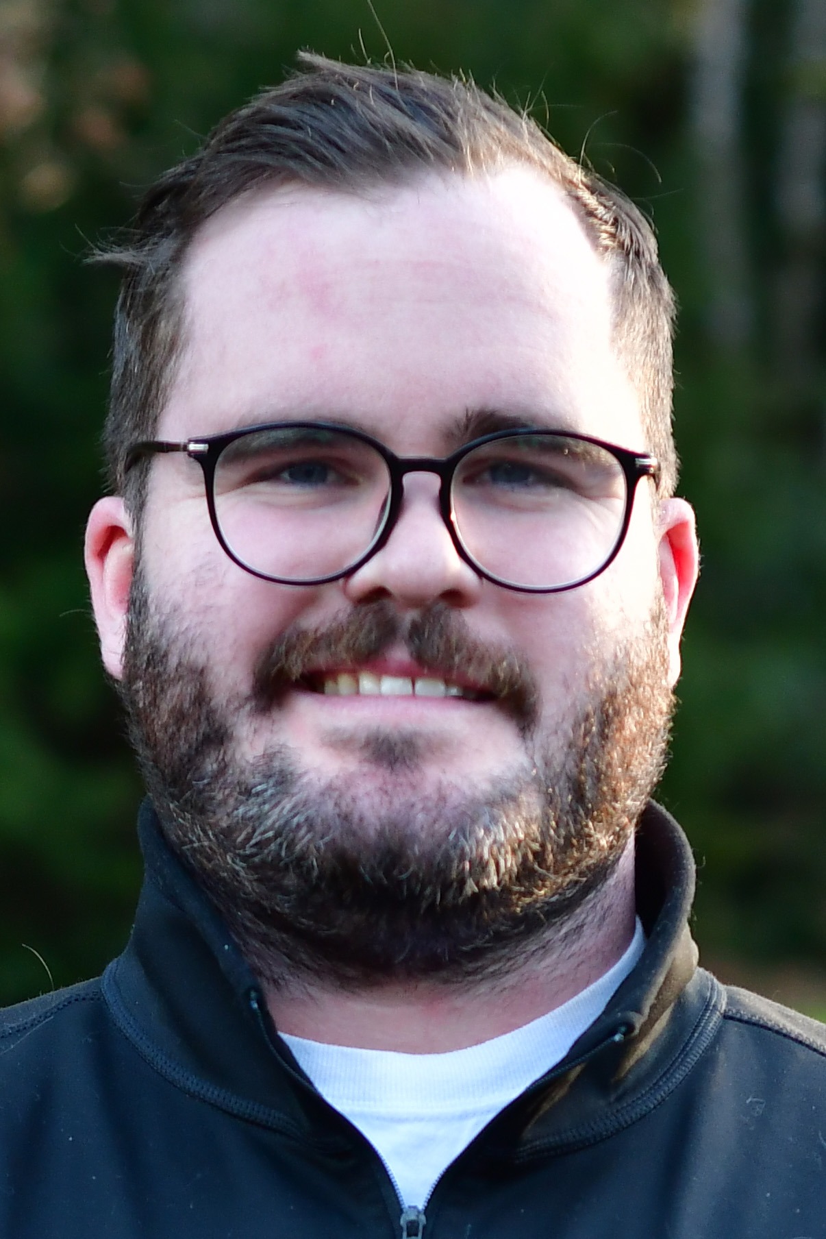 Adam Riddle stands outside in front of a green background, smiling.