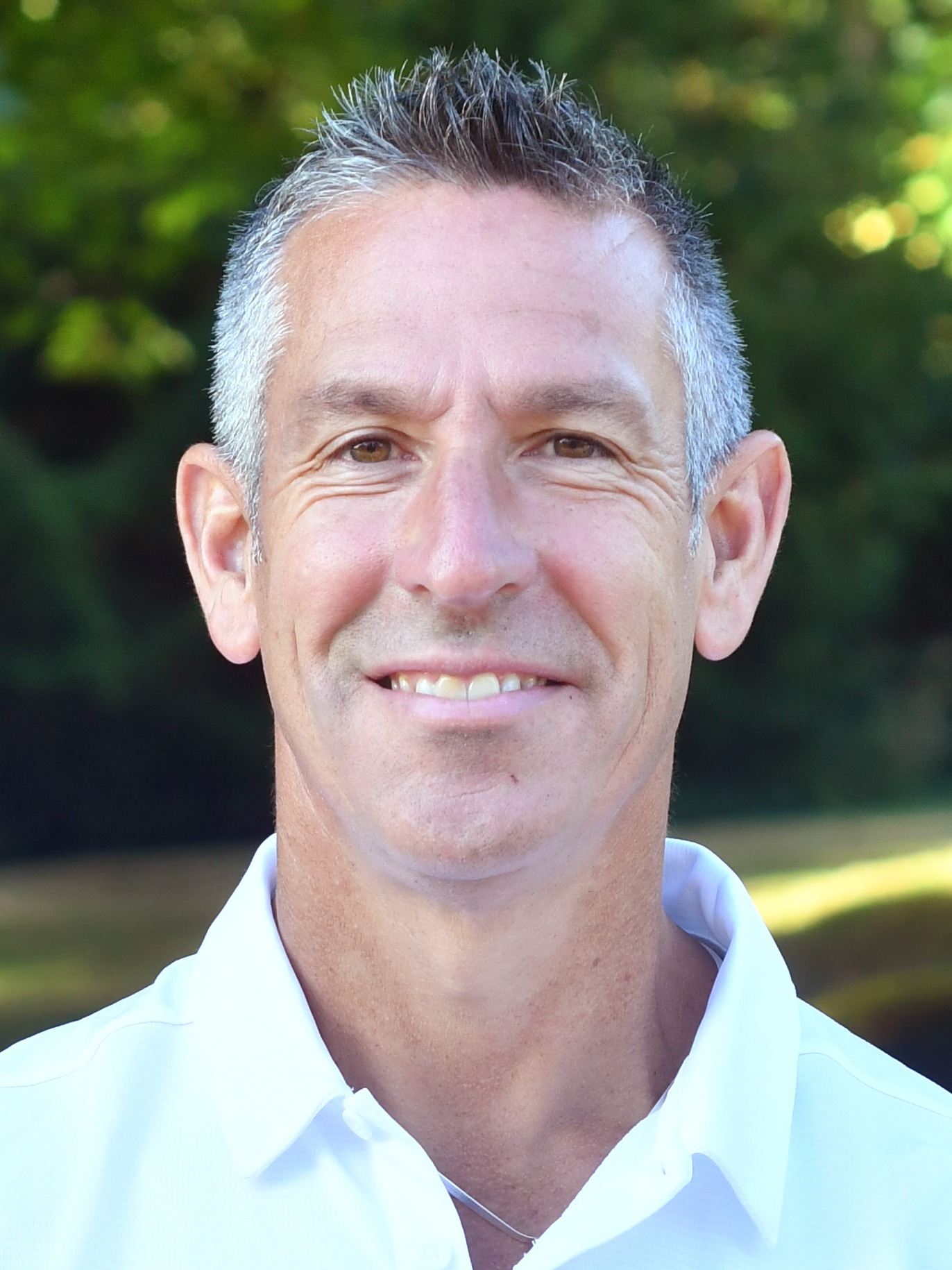 Joe Bushman stands outside in front of a green background, smiling.