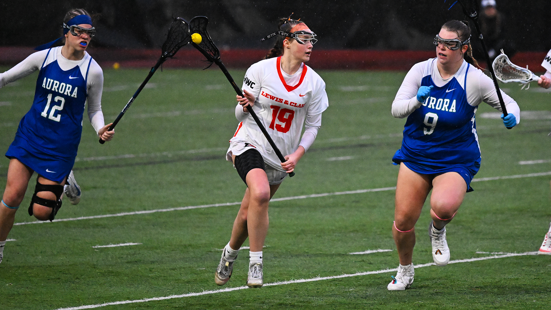 Kaedance Stubbs in a white uniform runs down the field with the ball between two defenders in blue uniforms. 