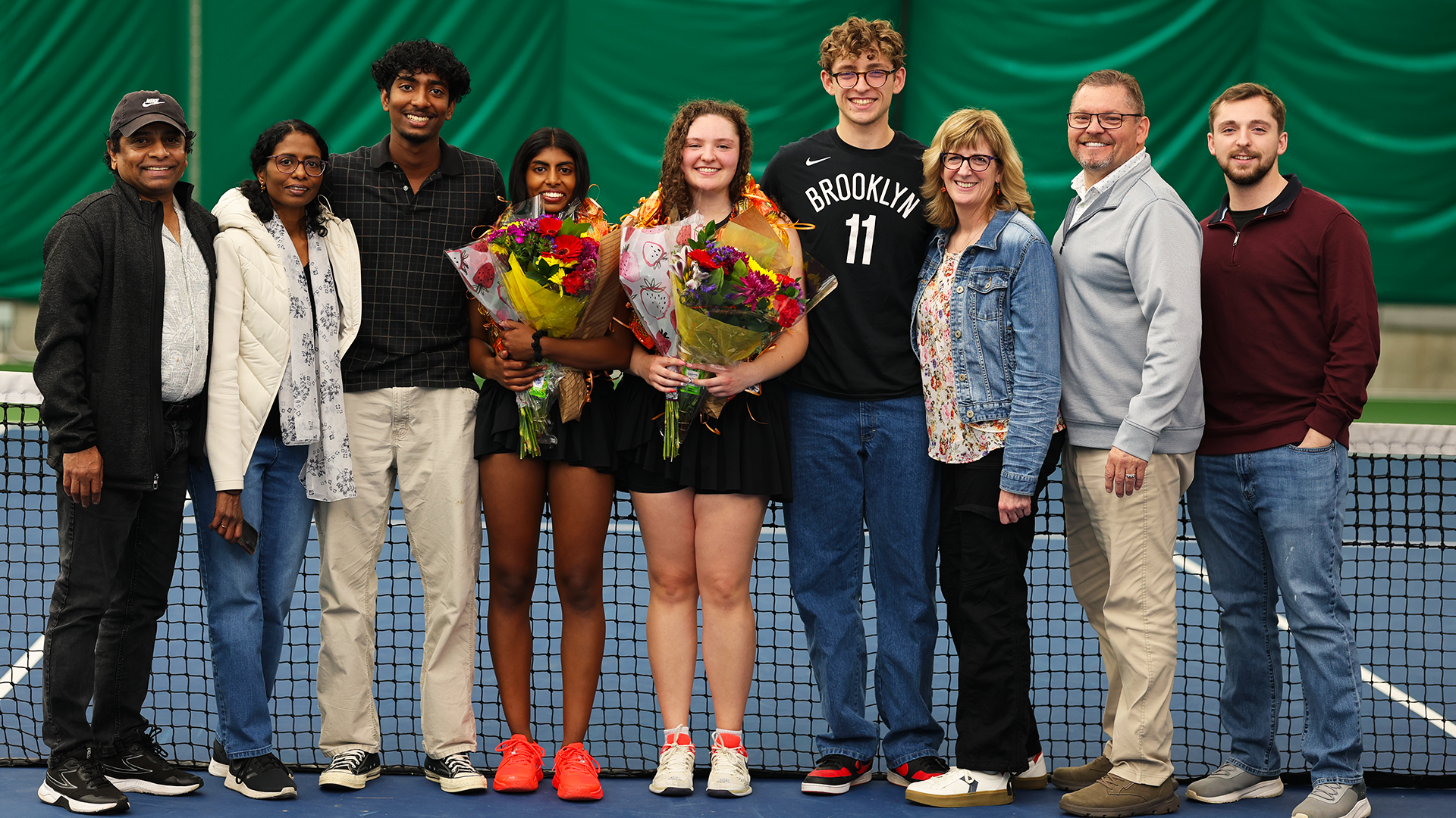 Women's Tennis seniors Susanna Anand and Emma Loucks pose with their families in front of a tennis net. 