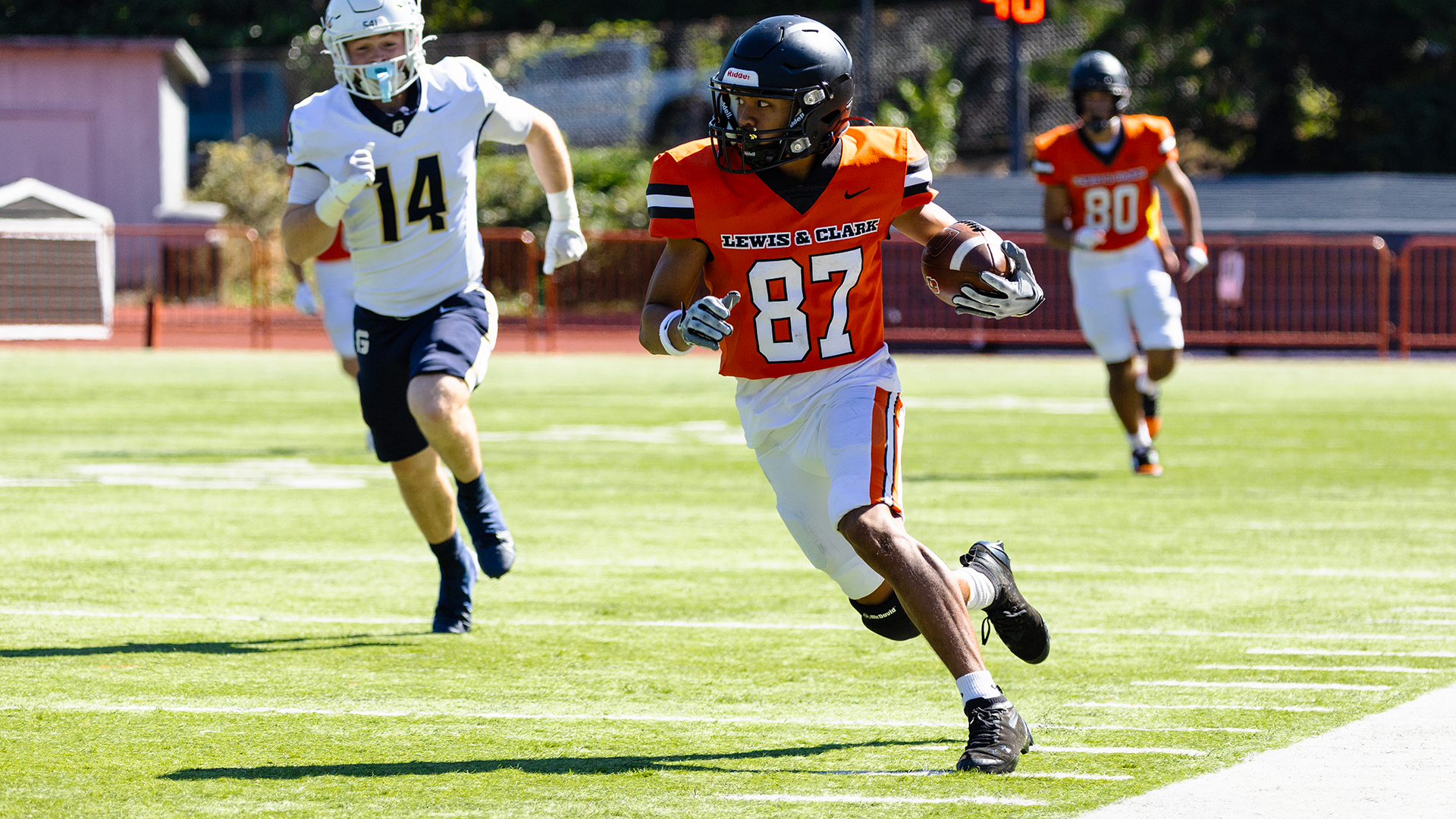 Photo of Sytyn Lasconia running with the football in his left arm wearing an orange jersey with white numbers, white pants and a black helmet with a player in a white jersey running after.