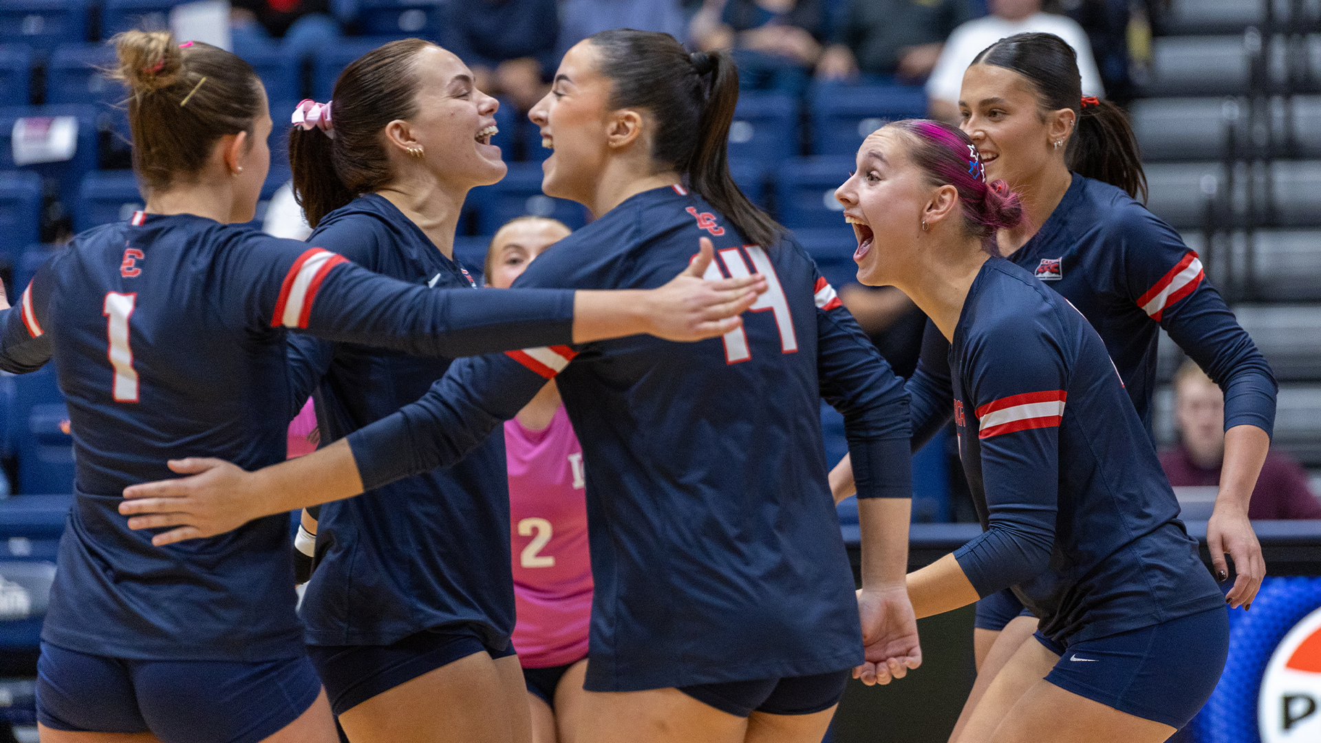 LC State Volleyball players in navy jerseys gather and celebrate on the court during a home match, with a player in a pink libero jersey visible in the center and spectators seated in the background.
