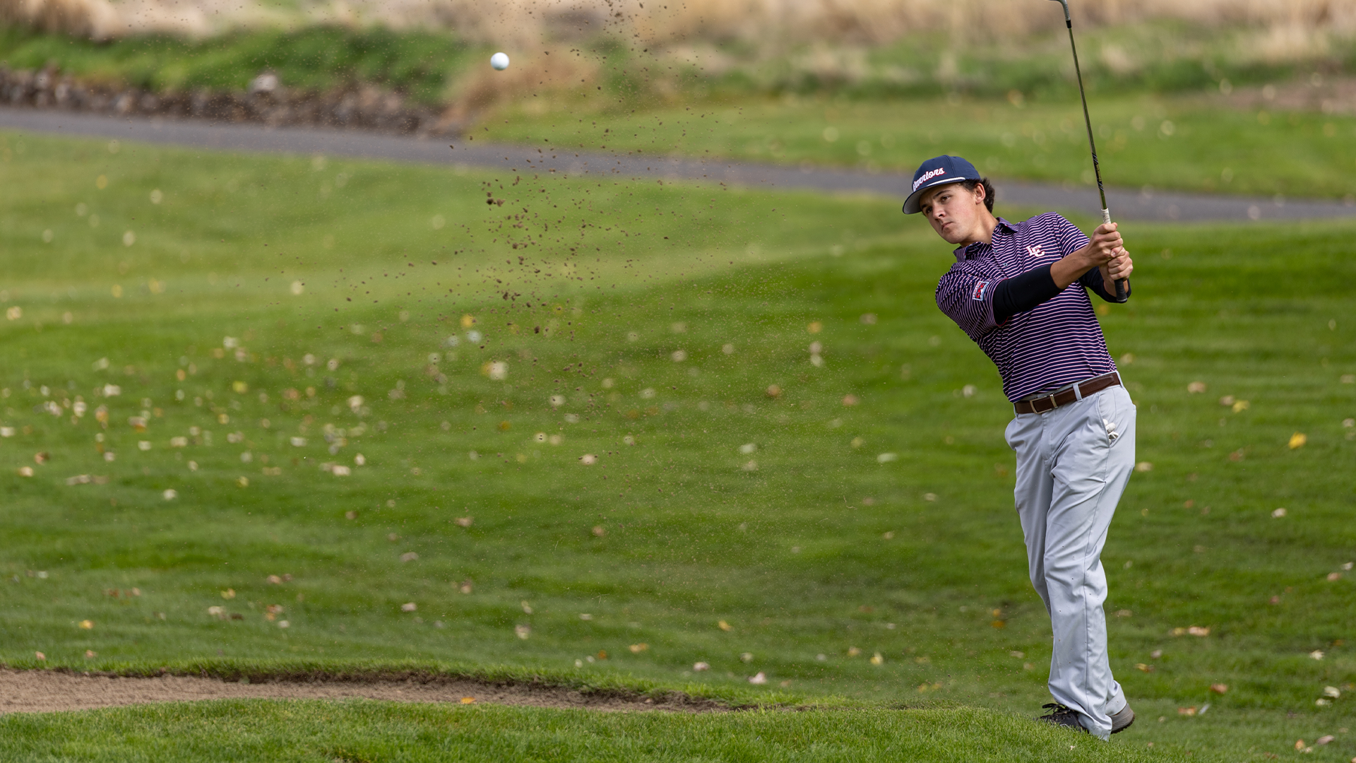 LC State men’s golfer hits an approach shot from a bunker, sending sand into the air as the golf ball travels toward the green on a tree-lined course.