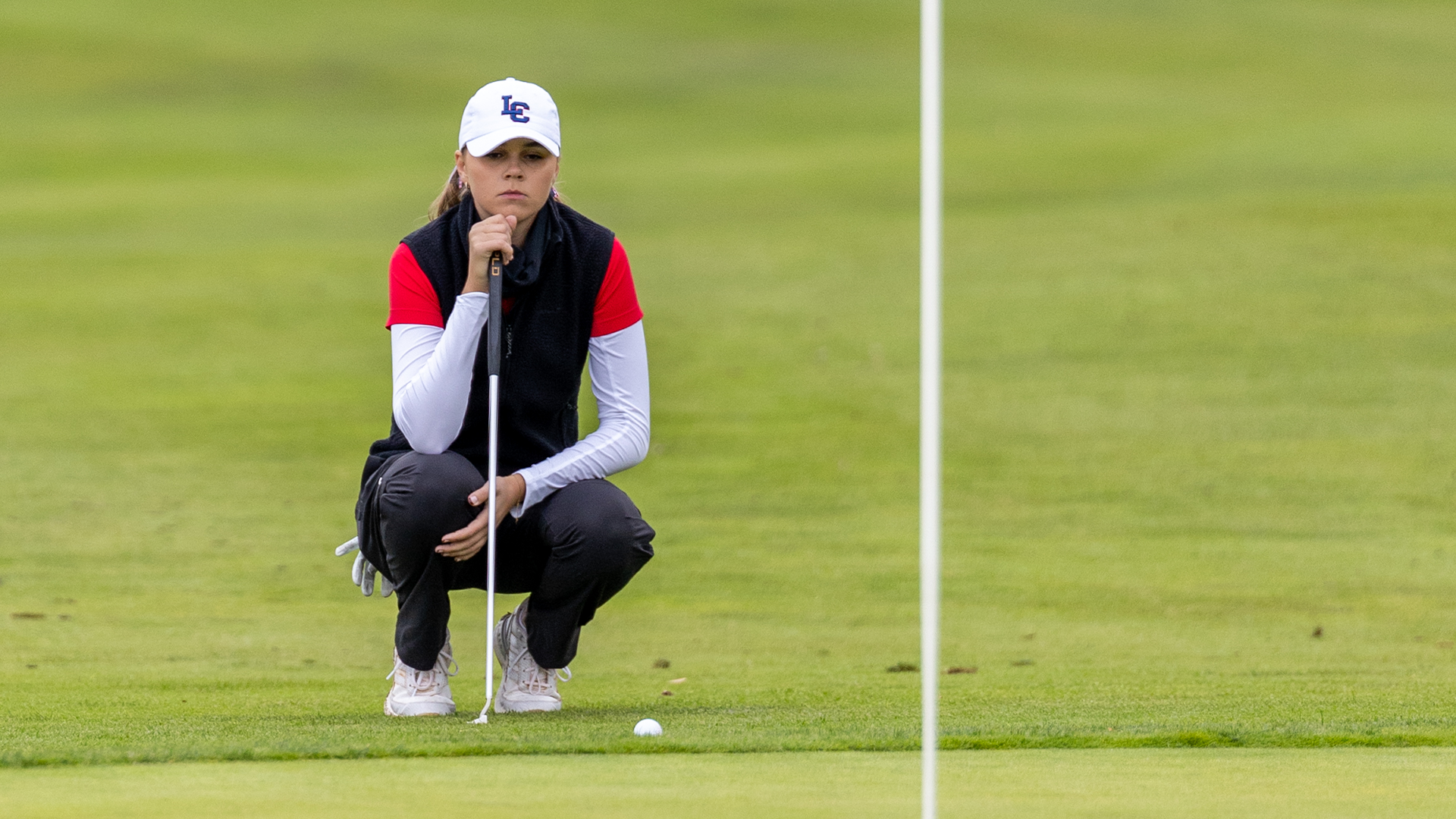 LC State women’s golfer crouches on the green and studies a putt with a white flagstick visible in the foreground and a golf ball resting between the golfer and the hole.