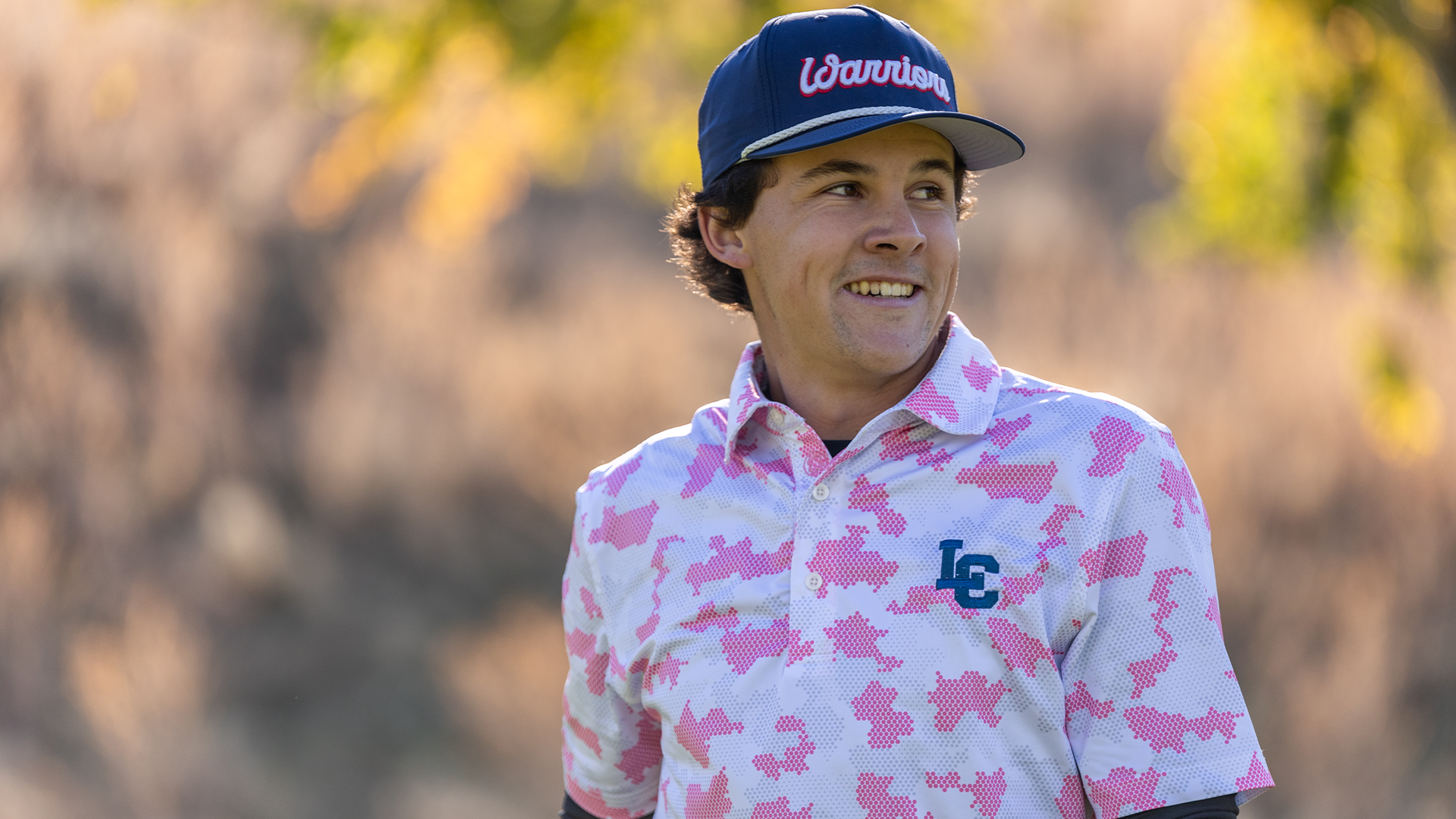 LC State men’s golfer wearing a pink breast cancer awareness polo stands on the course during competition, with autumn-colored trees softly blurred in the background.