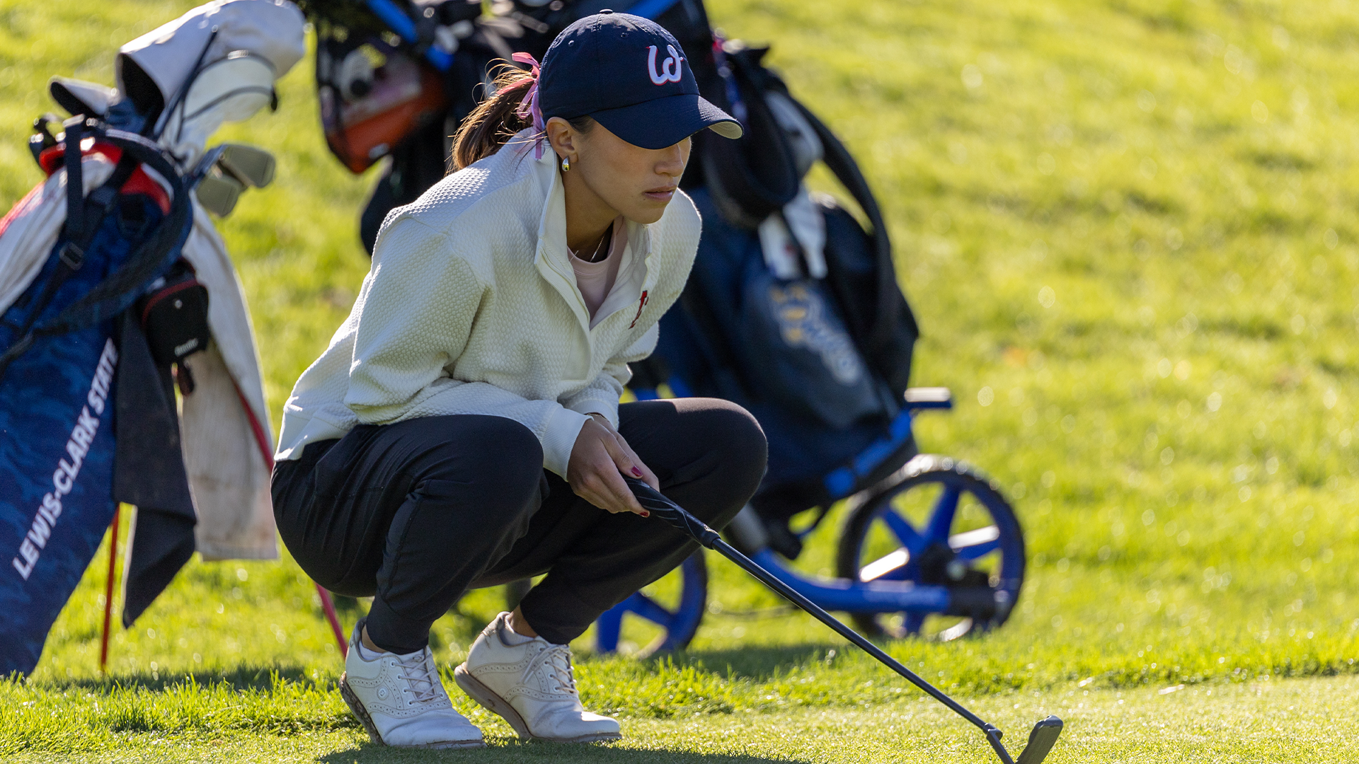 LC State women’s golfer crouches beside the green, lining up a putt with a club in hand while golf bags and push carts sit in the background on a sunlit course.