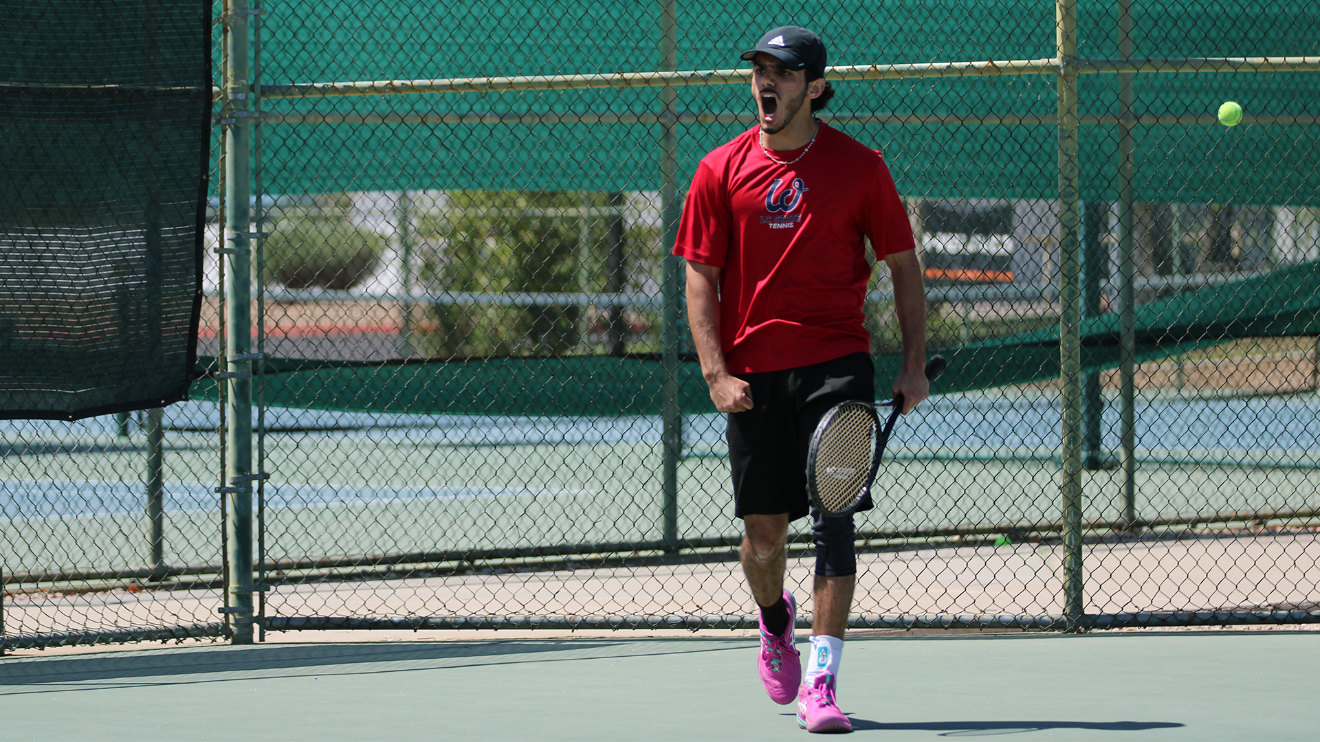 LC State men’s tennis player celebrates a point during a match, holding a racket and moving across the court as a tennis ball hangs in the air near the baseline.
