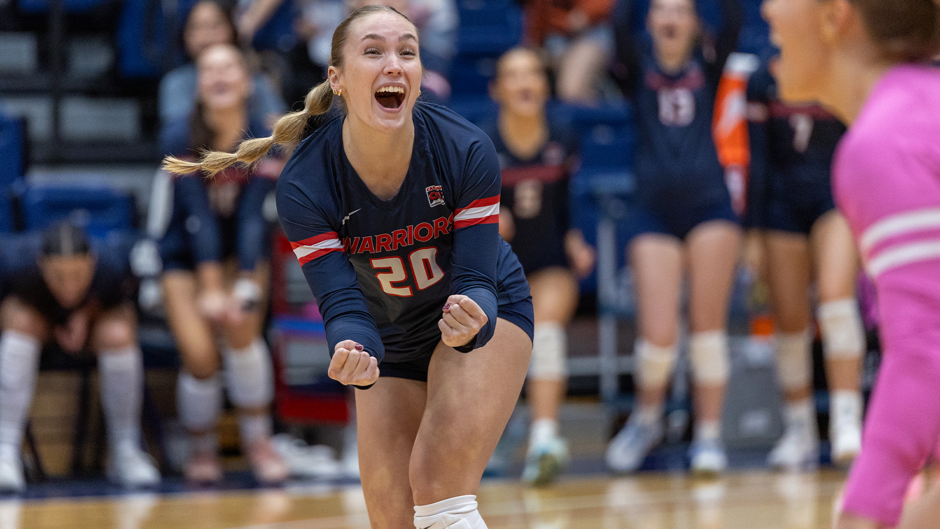 LC State Volleyball player in a navy Warriors jersey celebrates a point on the court during a home match, with teammates and spectators visible in the background