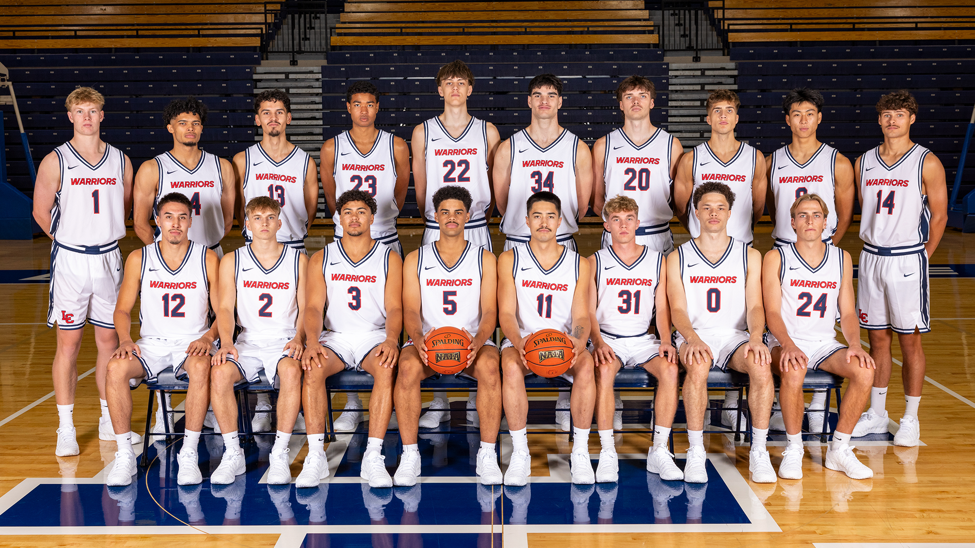 Official LC State Men’s Basketball team photo featuring players in white Warriors uniforms posed on the court inside the P1FCU Activity Center, with one row standing and one row seated in front of the arena bleachers.