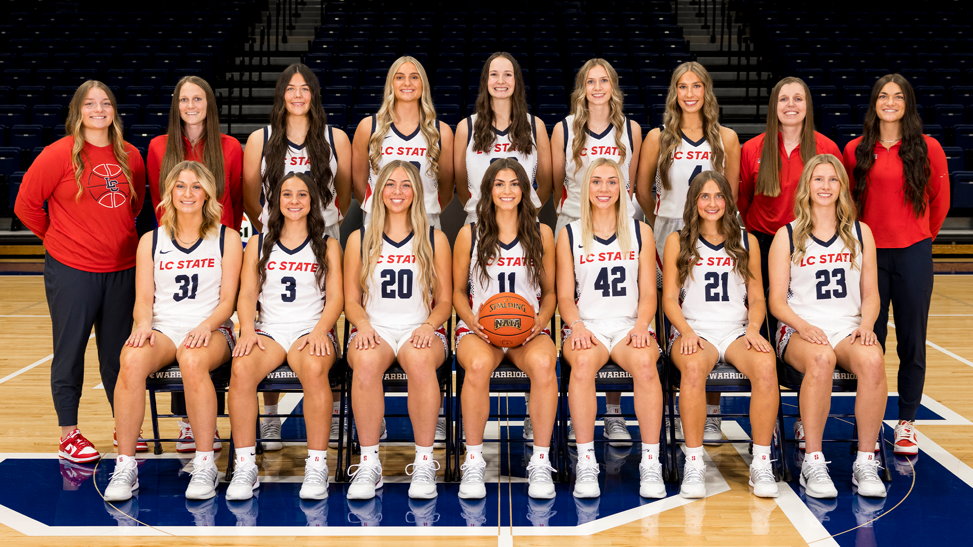 Official LC State Women’s Basketball team photo featuring players and coaches posed on the court inside the P1FCU Activity Center, with athletes in white game uniforms seated and standing in rows in front of the arena bleachers.
