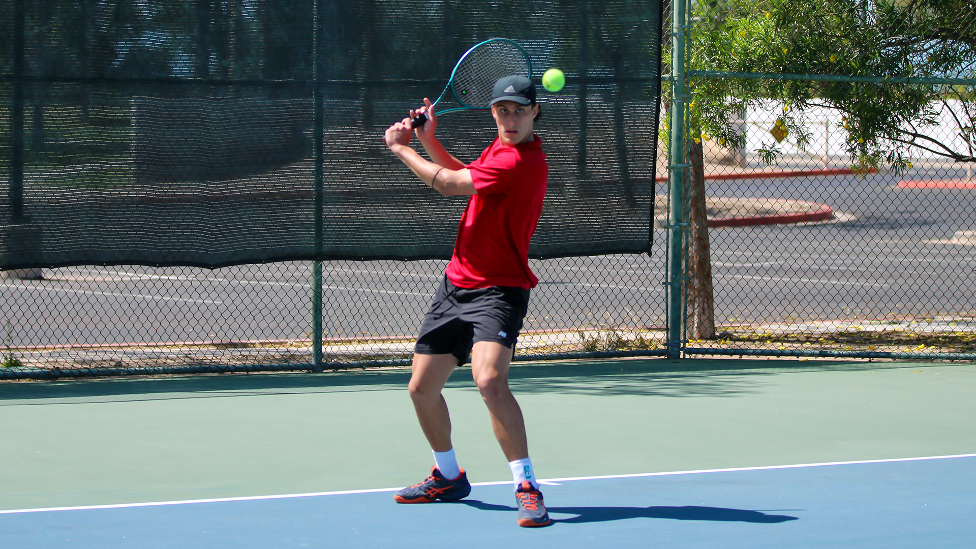 LC State men’s tennis player prepares to hit a two-handed backhand during a match, tracking a tennis ball in midair on an outdoor court with fencing and trees in the background.