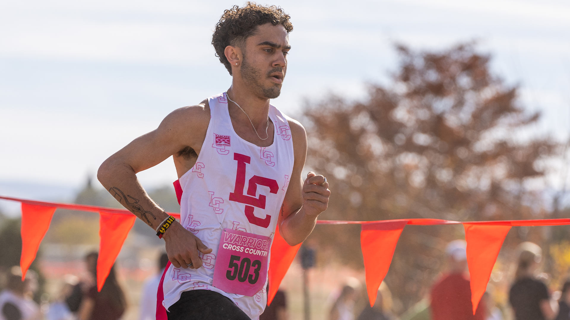 LC State men’s cross country runner races along a course lined with orange flags, wearing a white Warriors singlet with a pink race bib during a breast cancer awareness meet, with spectators and autumn trees blurred in the background.