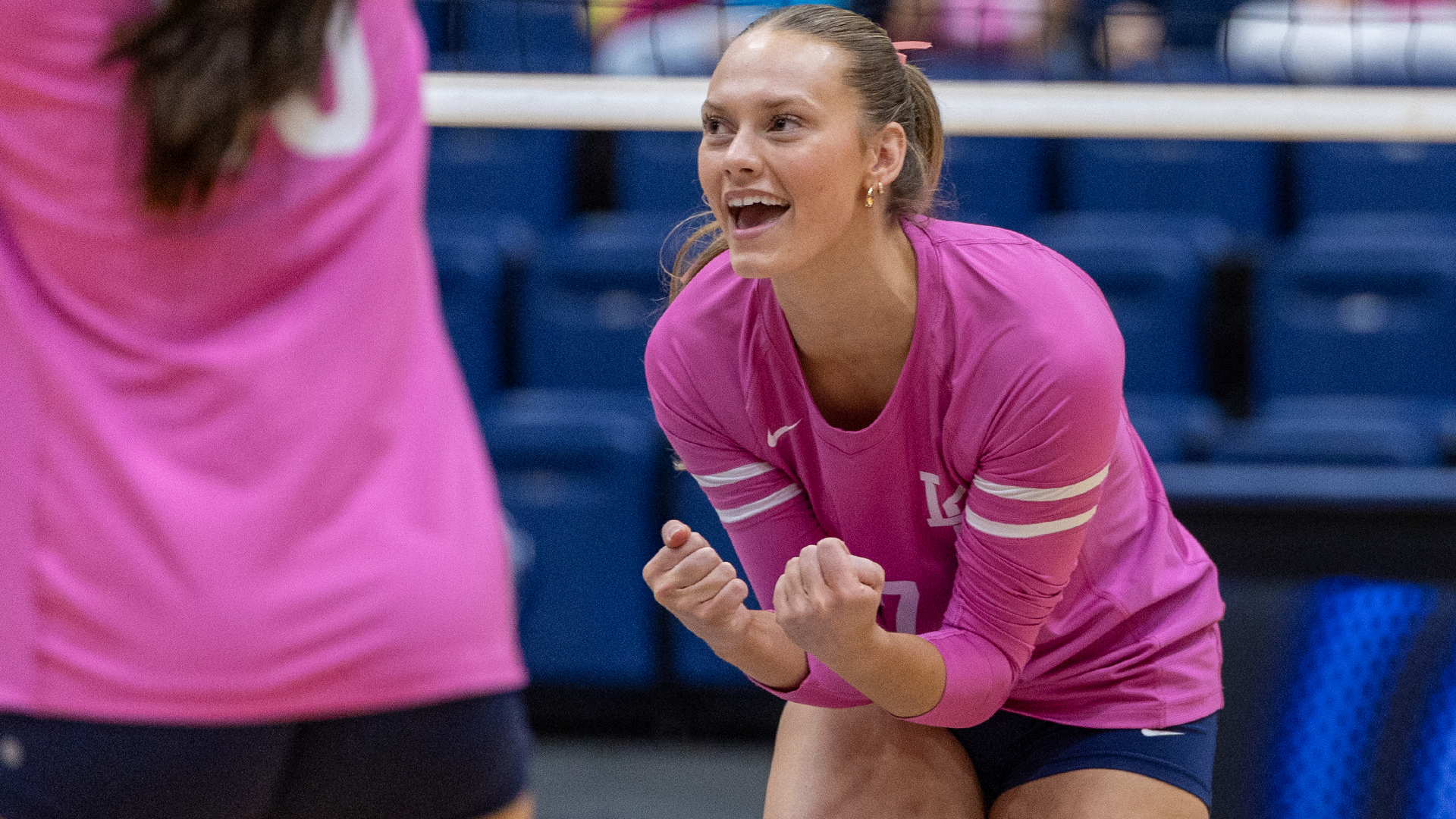 LC State Volleyball player in a pink jersey celebrates a point during Pink Night, crouched in a ready position on the court with clenched fists, while a teammate is visible in the foreground and blue arena seating appears in the background.