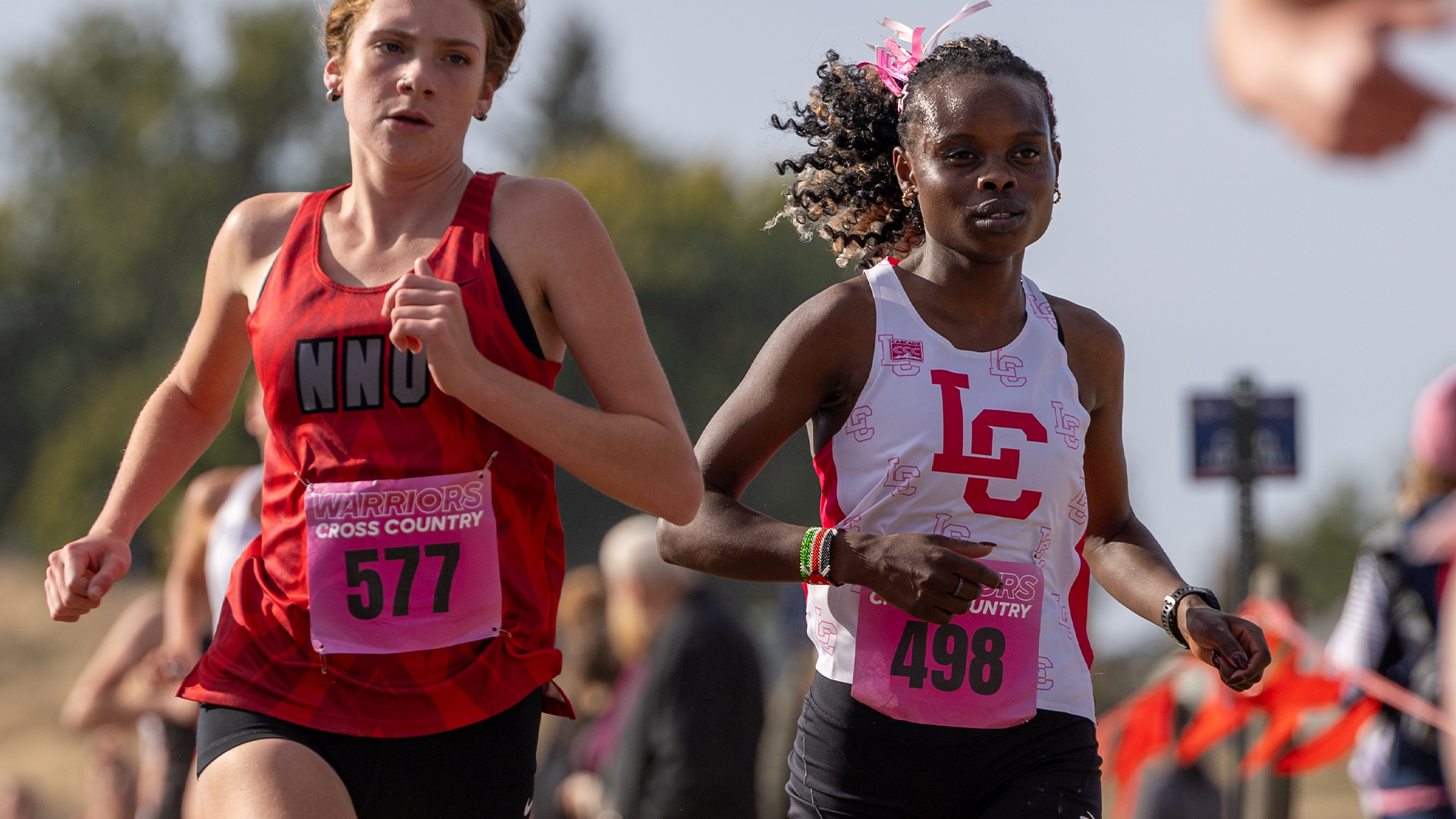 LC State women’s cross country runner competes alongside an opponent during a race, with both athletes wearing pink race bibs and breast cancer awareness accents as spectators and course markers blur in the background.