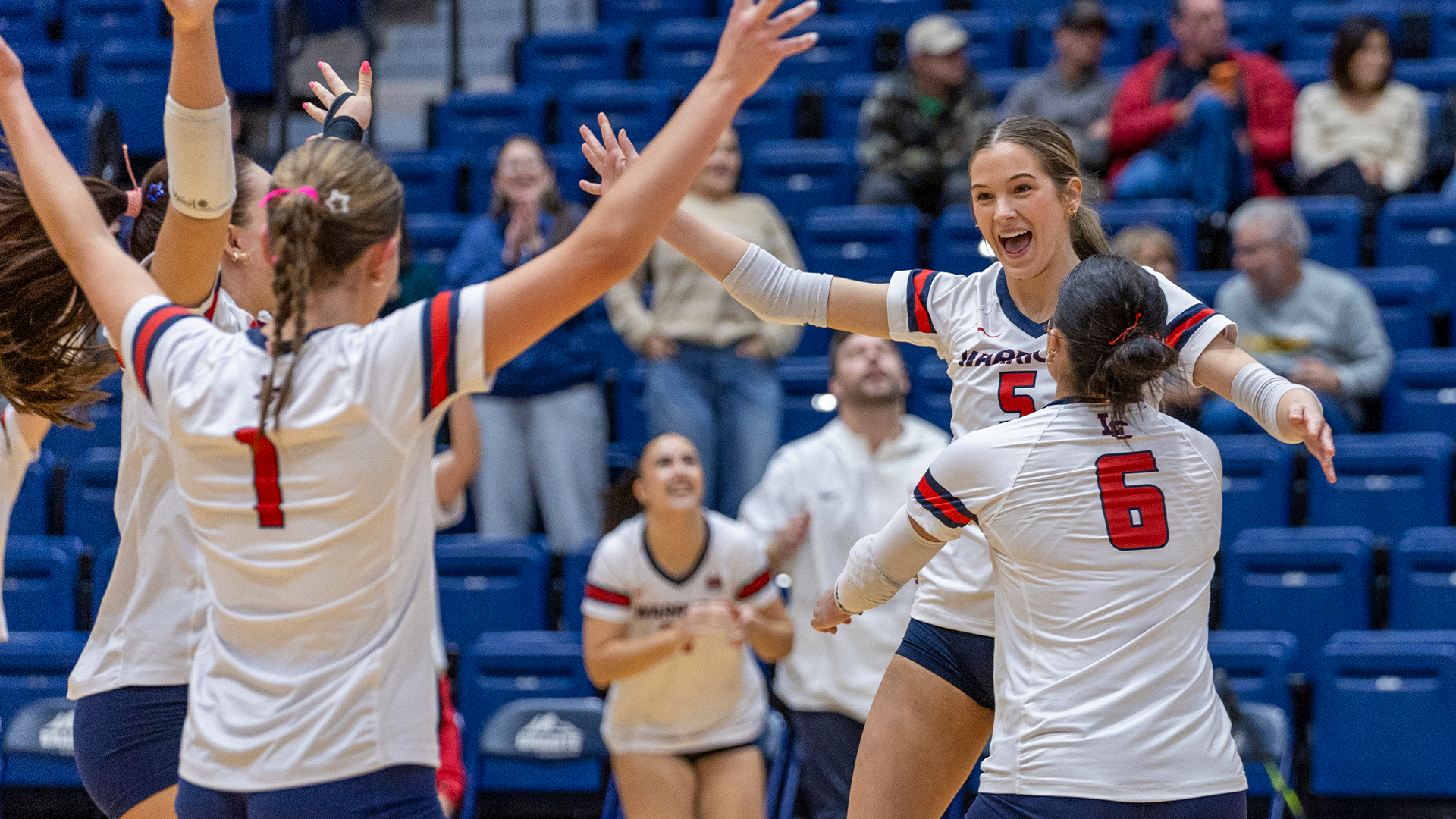 LC State Volleyball players in white jerseys celebrate a point on the court during a home match, with teammates gathered near the net, blue arena seating in the background, and members of the bench visible along the sideline.