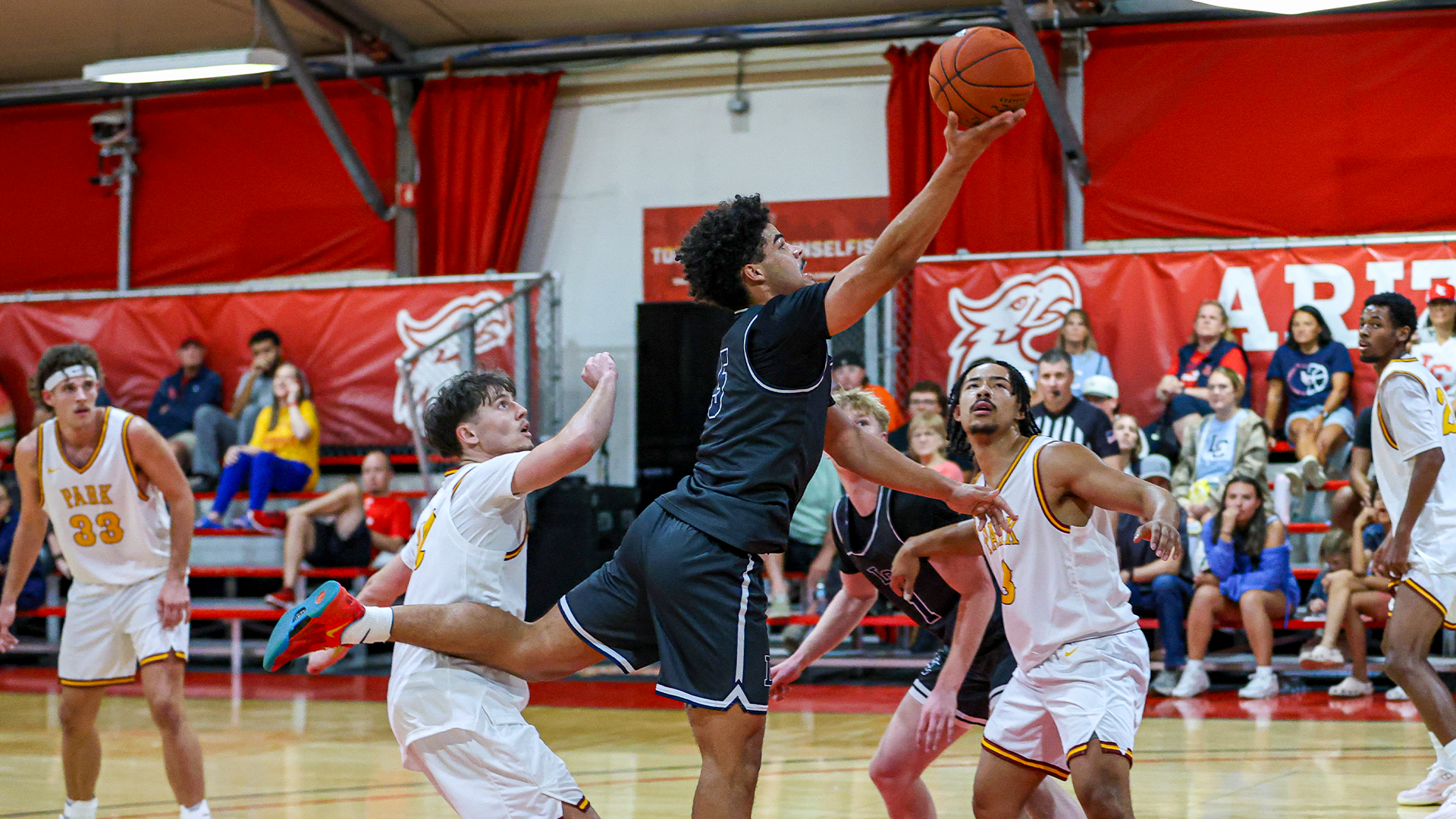 LC State men’s basketball player in a dark uniform drives to the basket for a layup between multiple defenders during a game, with spectators seated in red bleachers and players from both teams watching the play unfold.