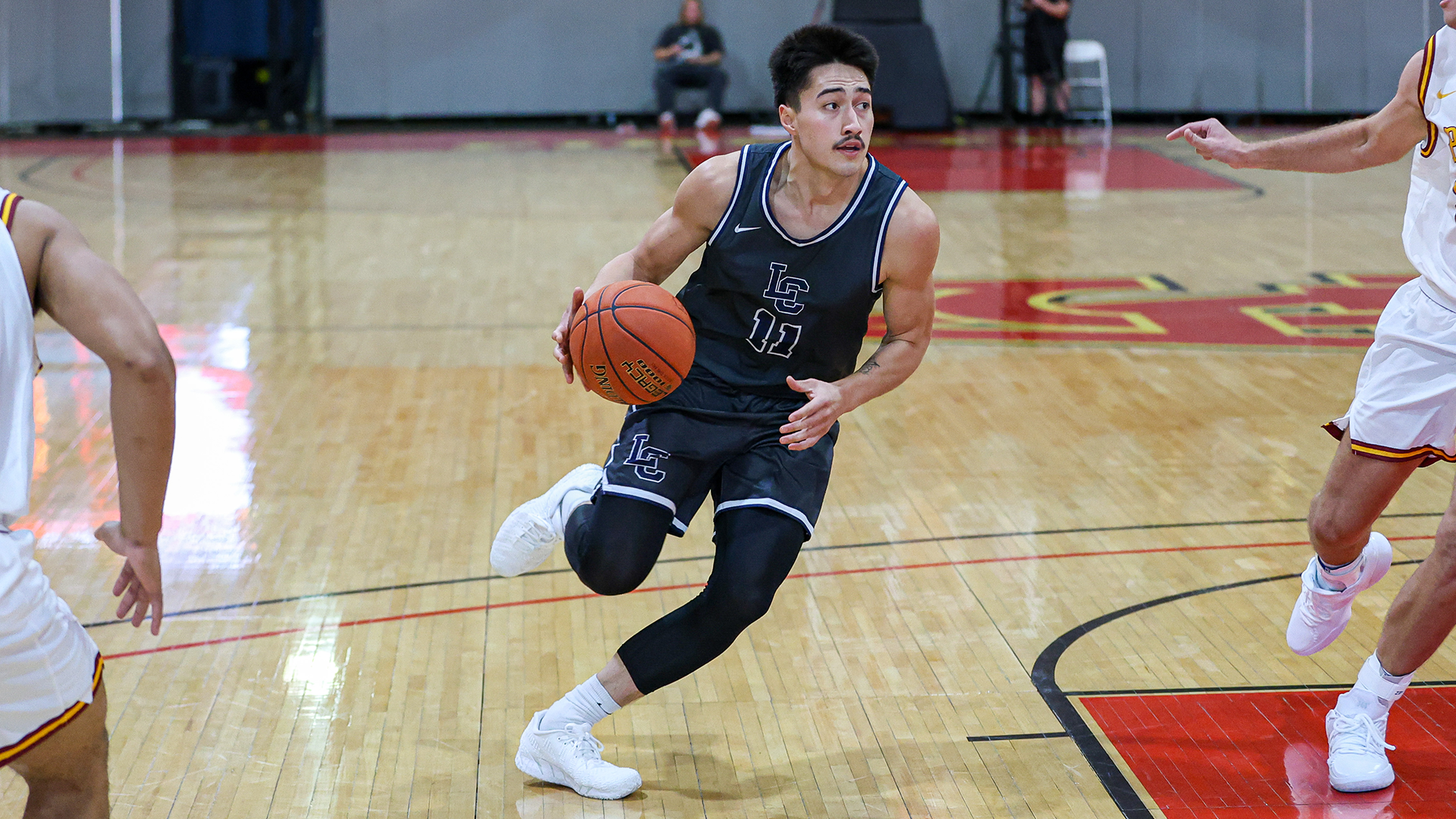 LC State men’s basketball player in a dark Warriors uniform dribbles between defenders during a game, driving toward the basket on a hardwood court while opposing players in white jerseys close in from both sides.