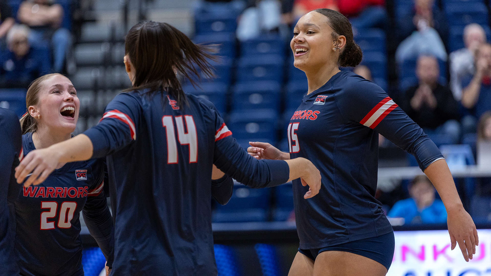 LC State Volleyball players in navy Warriors jerseys gather on the court during a home match, with multiple teammates visible near the net and blue arena seating in the background.
