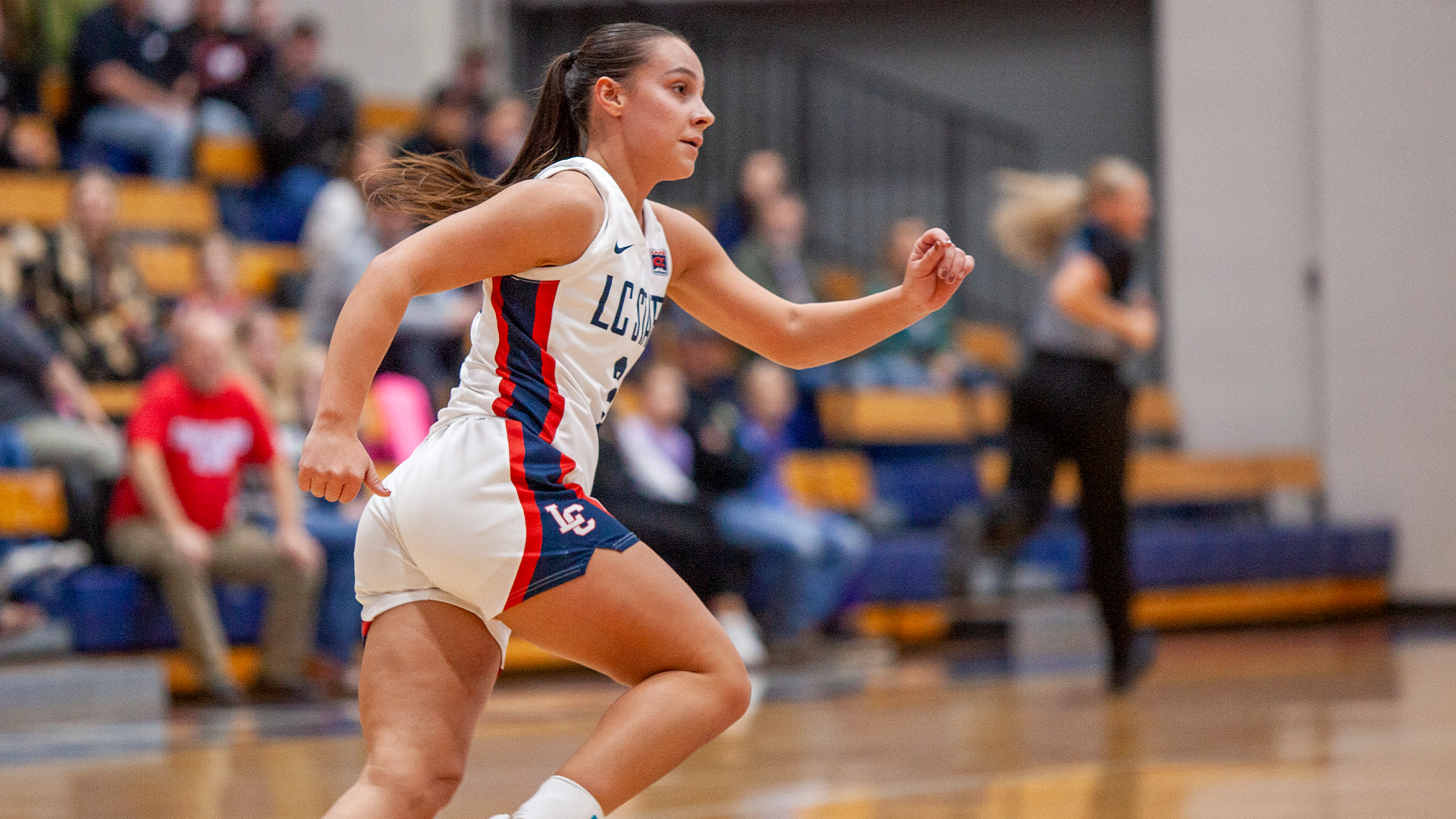 LC State women’s basketball player in a white Warriors uniform runs up the court during a game, with spectators seated in the bleachers and the hardwood court visible in the foreground.