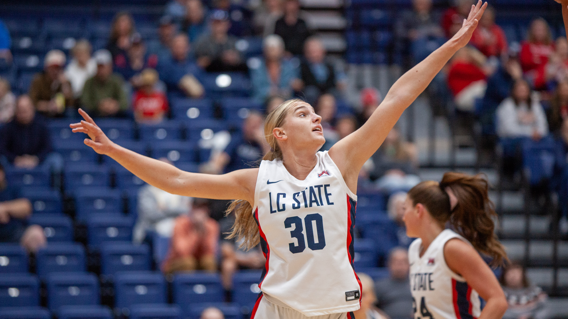 LC State women’s basketball player in a white Warriors uniform reaches upward to contest or receive the ball during a game, with blue arena seating and additional players visible in the background.