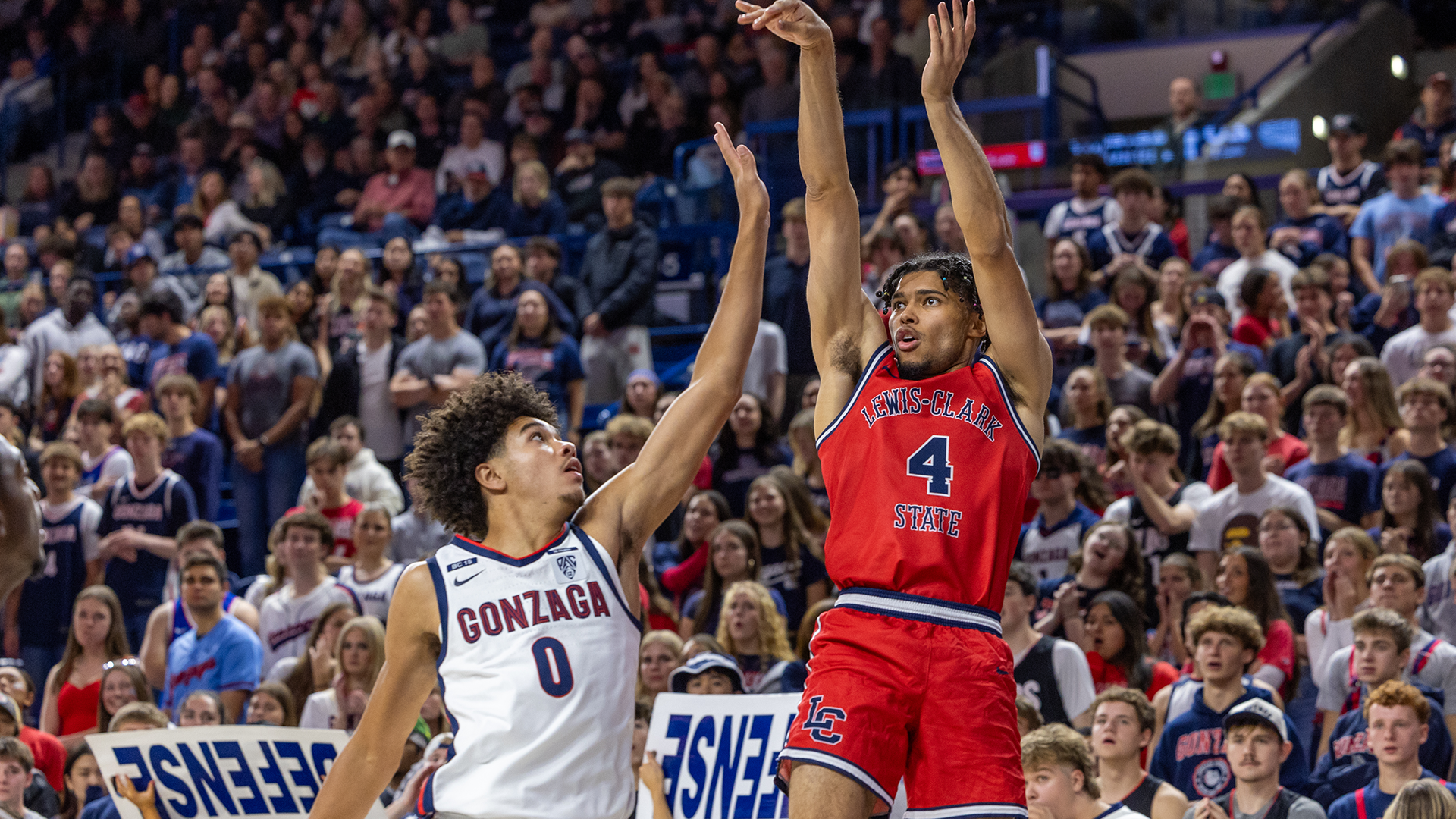 LC State men’s basketball player in a red Warriors uniform rises for a jump shot over a Gonzaga defender during a game at a packed arena, with a large crowd filling the stands in the background.
