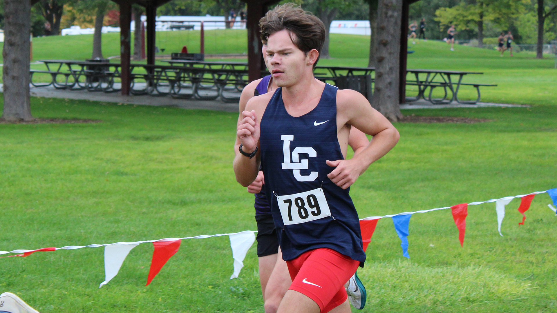 LC State cross country runner races along a grassy course marked with red, white and blue flags, with competitors and park shelters visible in the background.