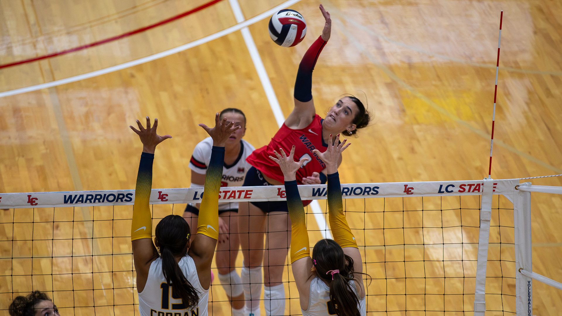 LC State Volleyball attacker reaches above the net for a spike during a match while two opposing blockers attempt to defend at the net.