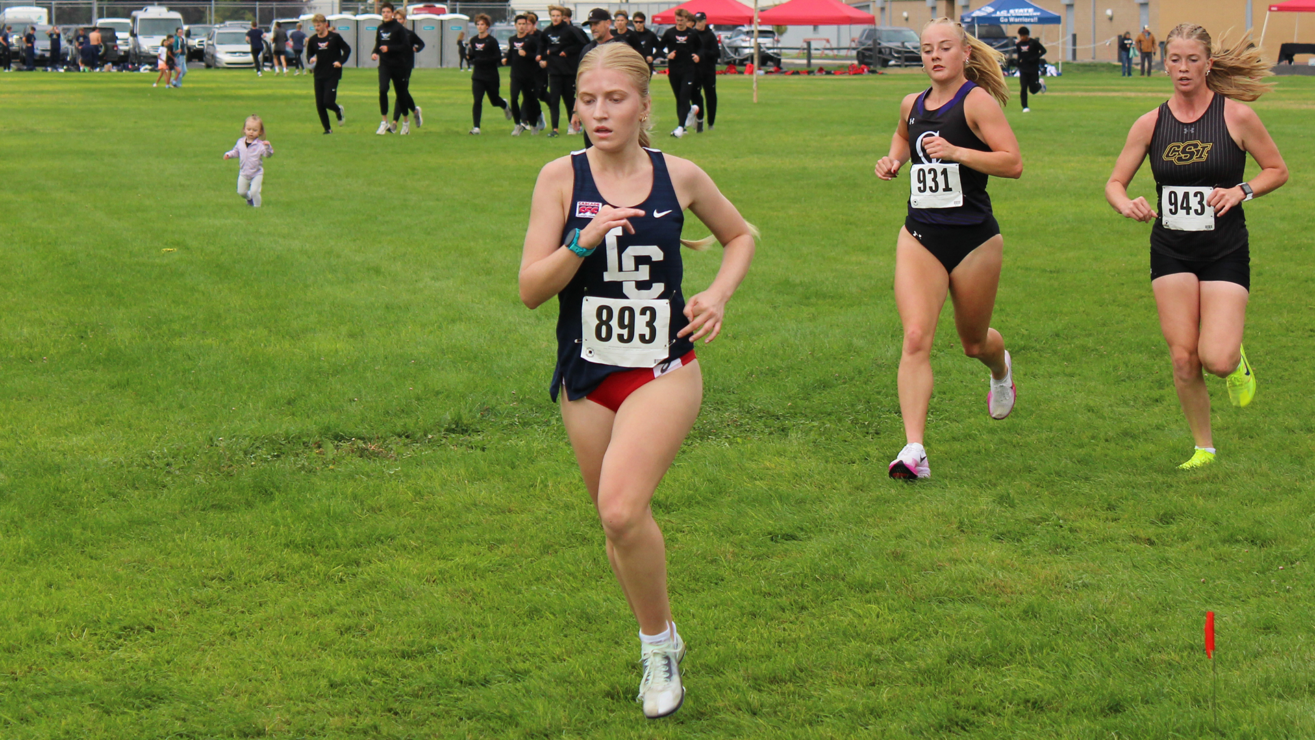 LC State cross country runner leads a pack of competitors through a grassy section of a race course, with other runners and spectators visible in the background.