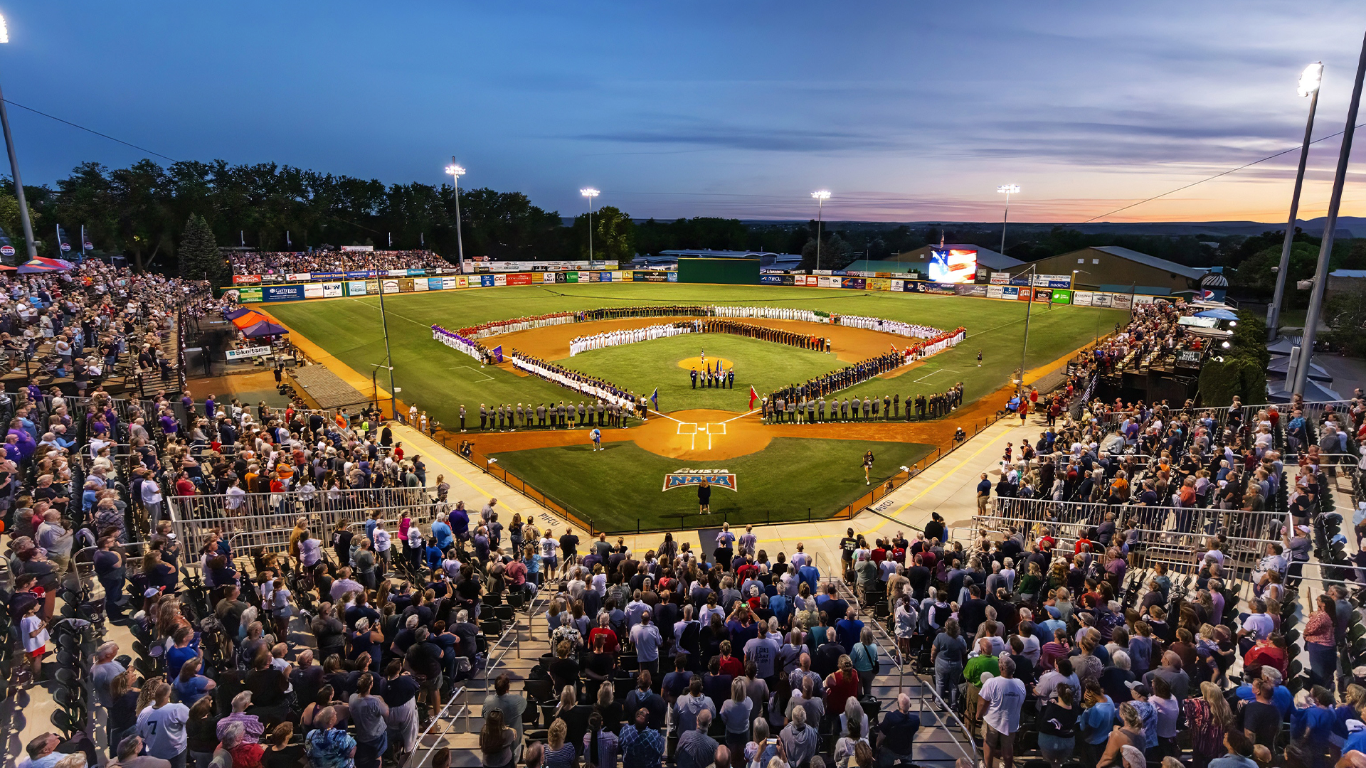 Wide view of a packed baseball stadium during an NAIA World Series opening ceremony, with teams lined up around the infield and spectators filling the stands at sunset.